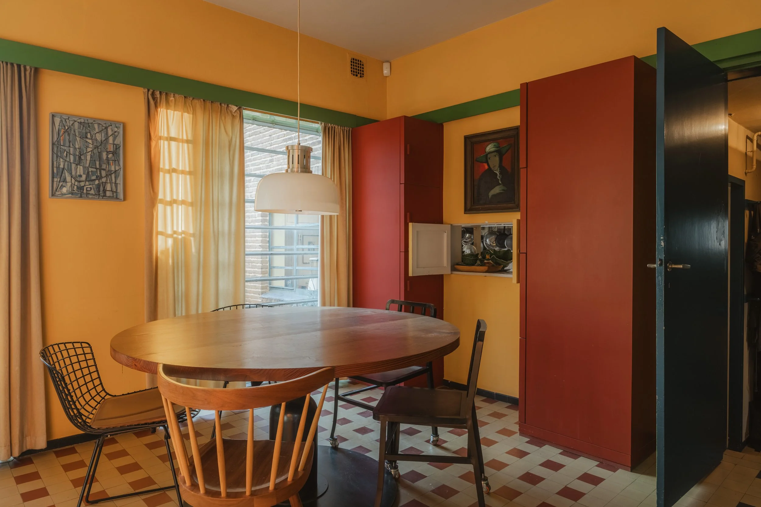 A dining room with yellow walls, a wooden oval table, and four chairs, two black wireframe, one wooden, and one dark brown. Beige curtains cover a window, with sunlight coming through. Red cabinets with built-in shelves and a painting of a woman wear