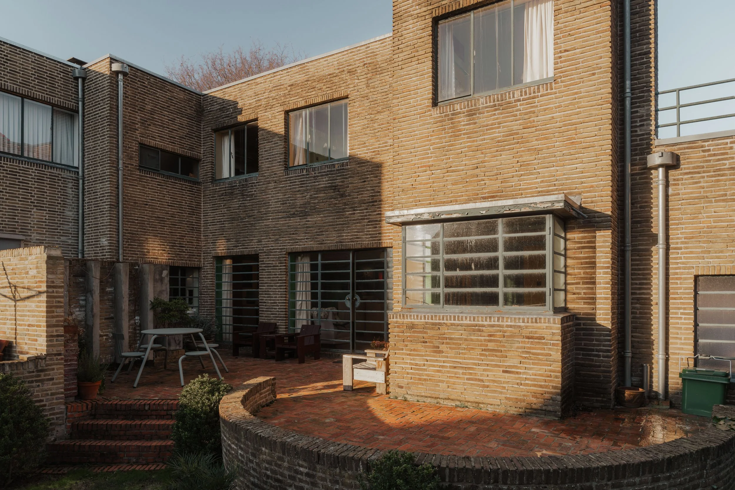 Brick building with multiple windows and a small outdoor patio area with chairs, table, and plants.