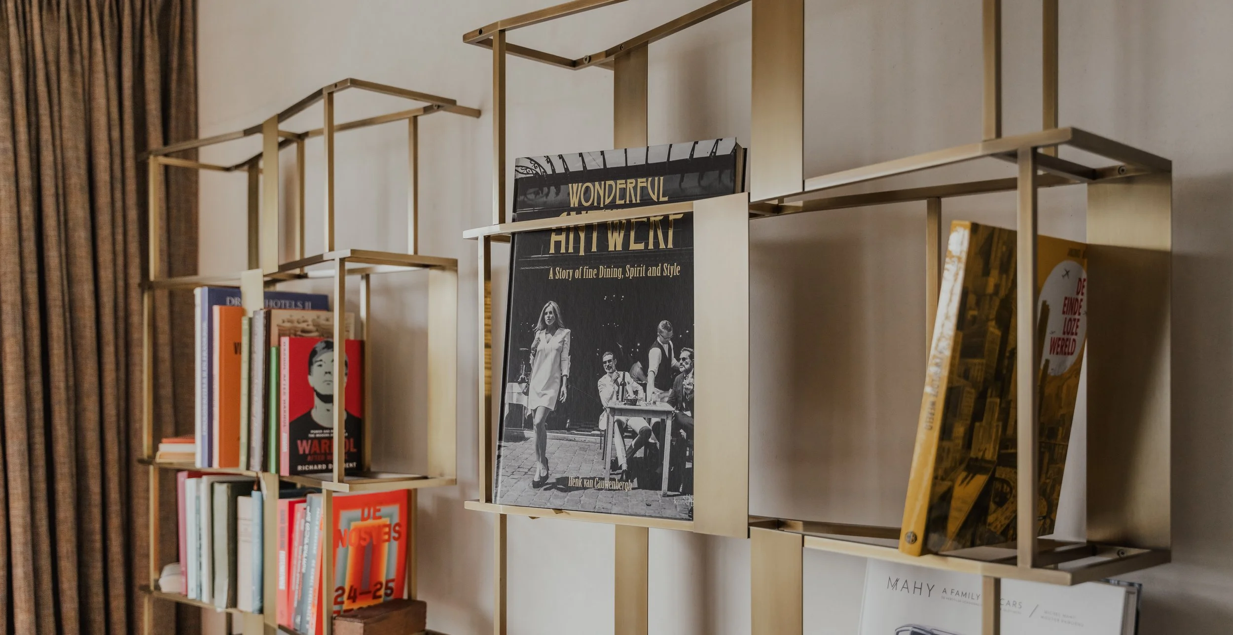 Books on a multi-level wooden bookshelf against a wall with a curtain on the left side.