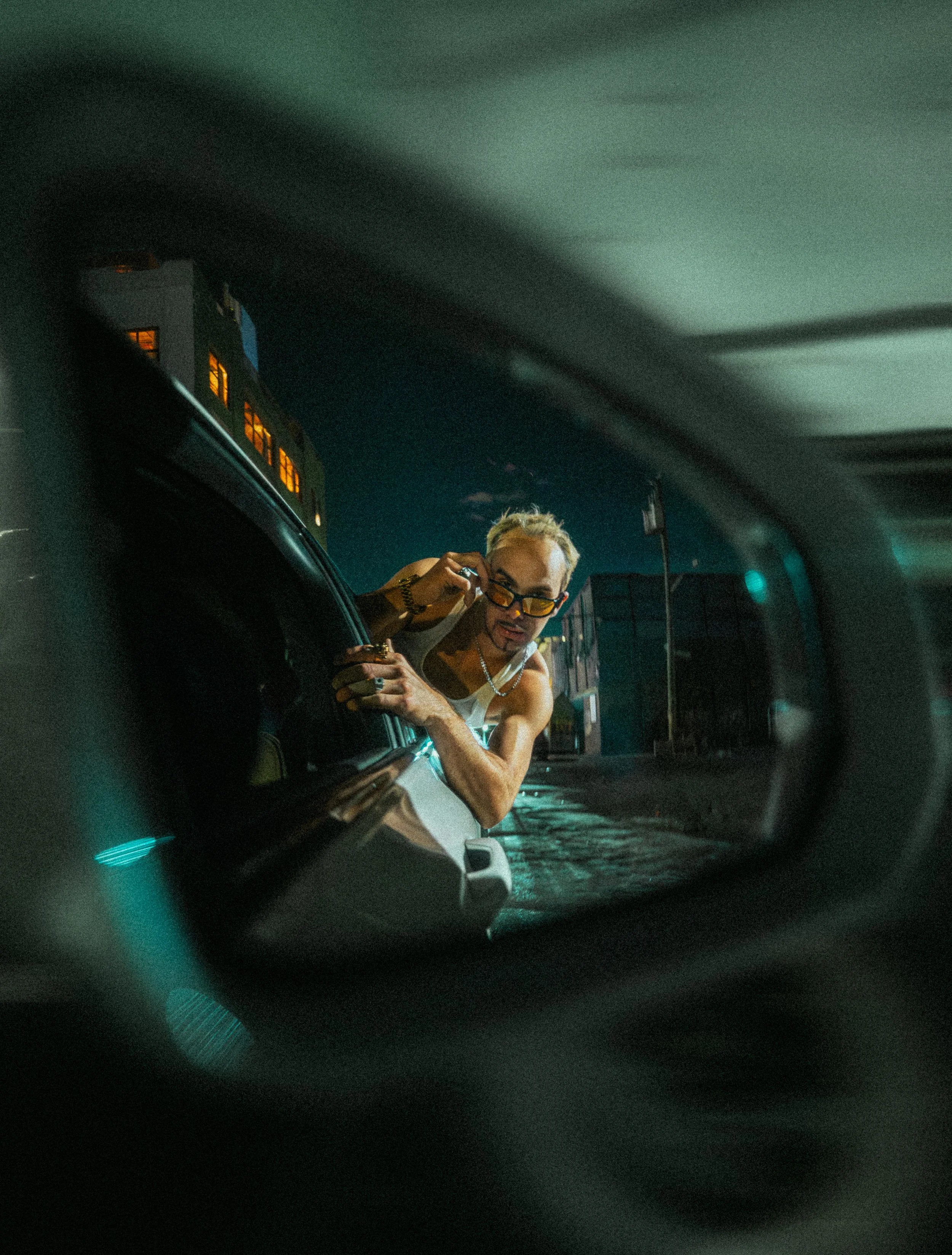A person leaning out of a car window, adjusting sunglasses, in a nighttime urban setting.