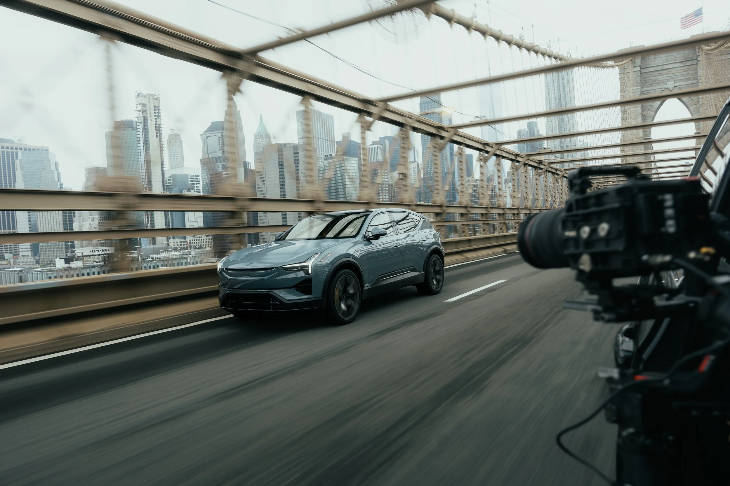 A gray electric SUV driving on the Brooklyn Bridge in New York City, with skyscrapers in the background and a camera mounted on a mechanical rig to the right.