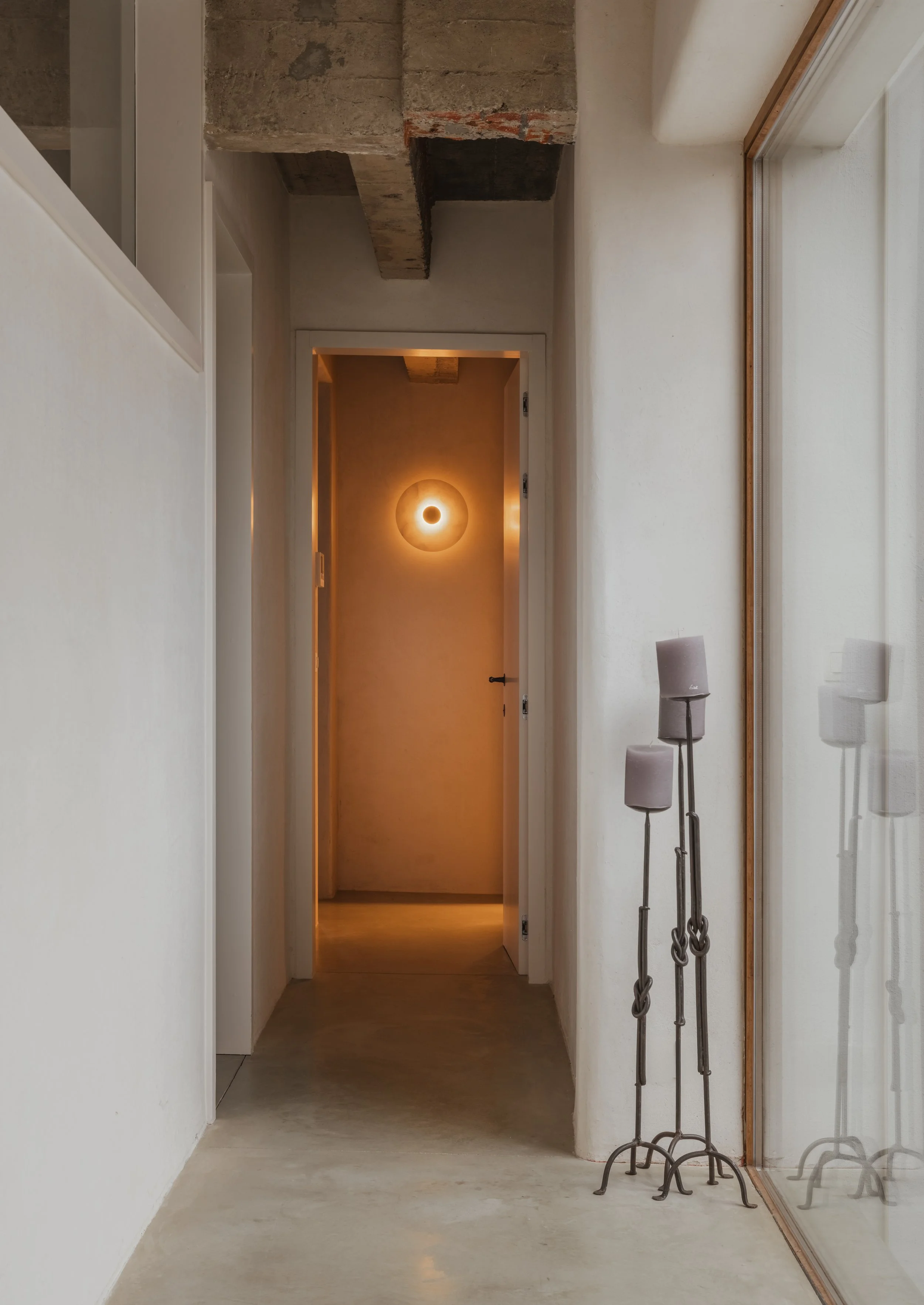 A minimalist hallway with white walls, concrete floor, and a reflective glass wall. There's a modern floor lamp with three knotted metal stands and fabric shades. An abstract round wall light is visible in the background. Exposed concrete ceiling bea