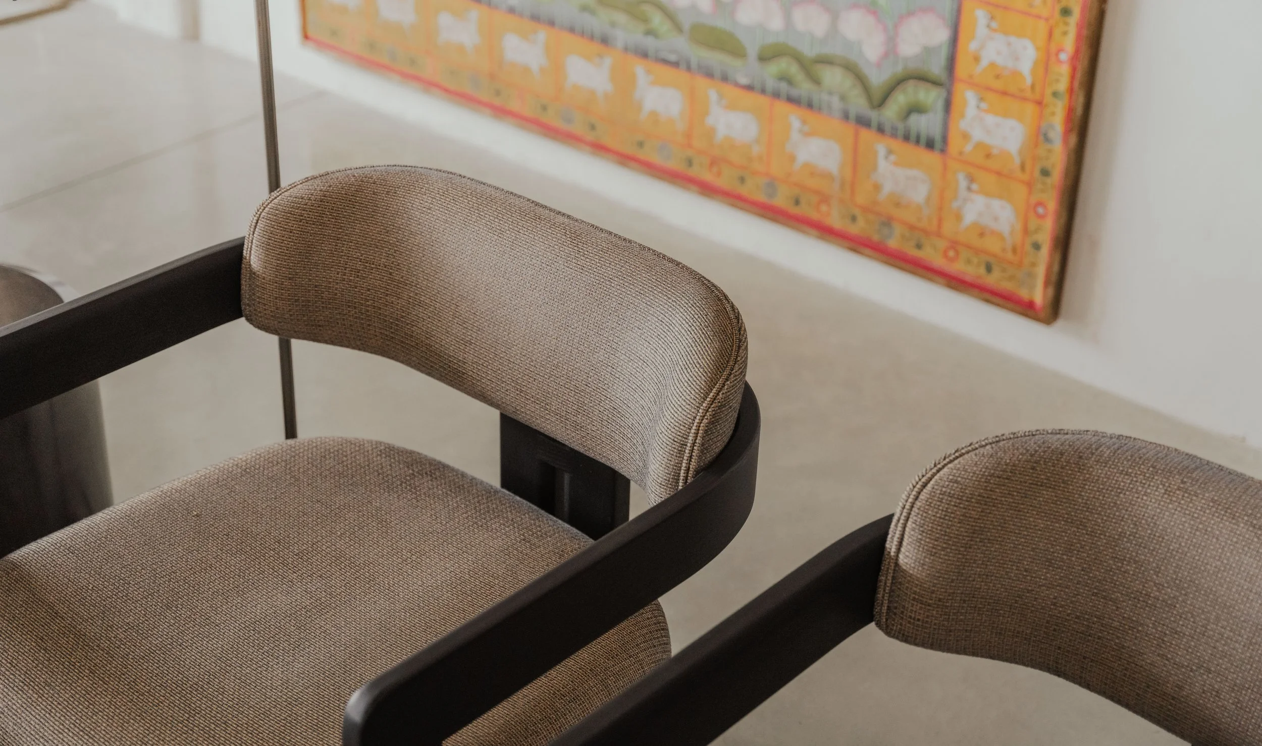 Close-up of two beige upholstered chairs with dark wooden frames in front of a colorful artwork on a white wall.