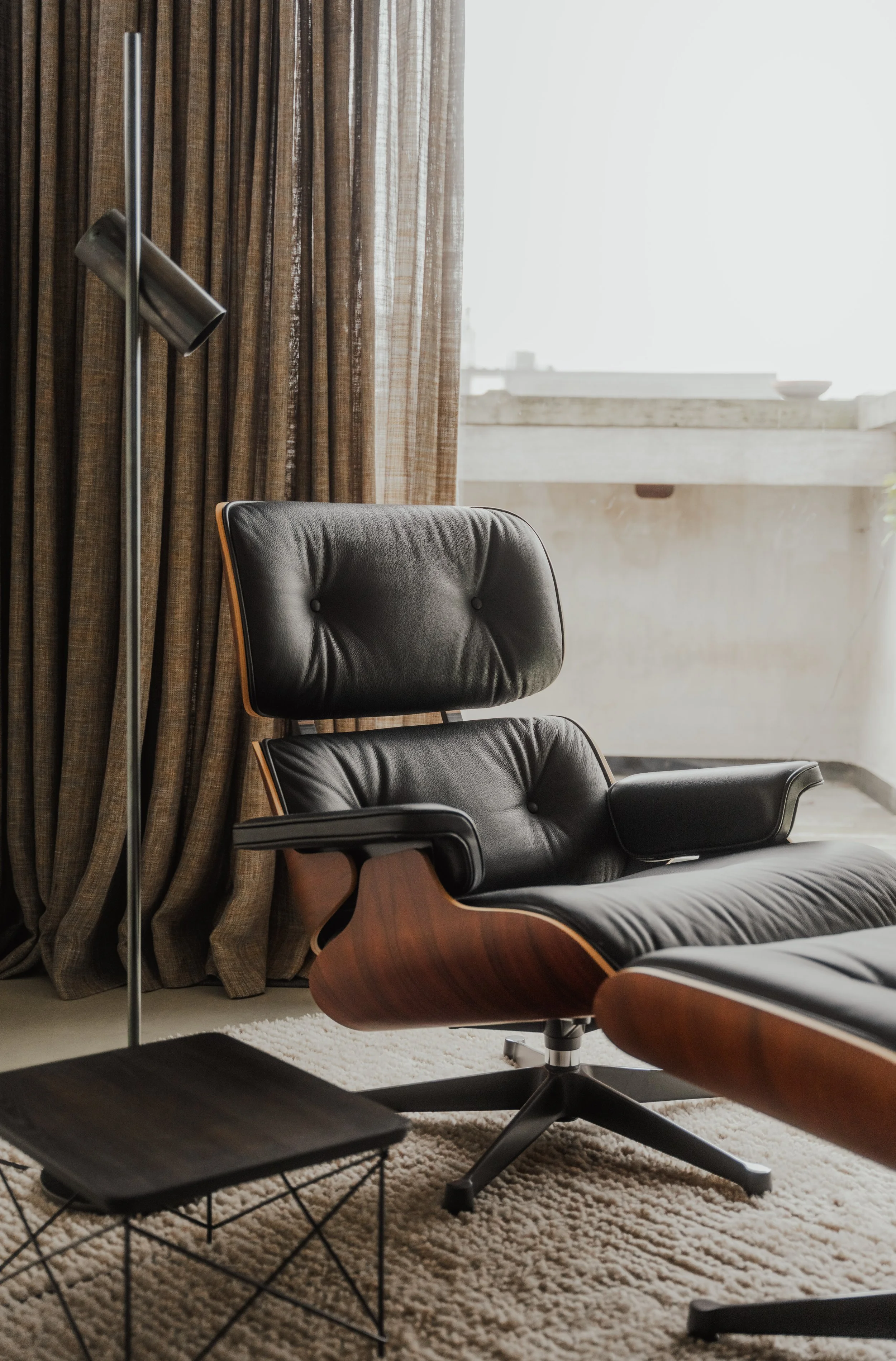 Modern black leather lounge chair with wooden accents, beige curtains, and a metal floor lamp next to a window with white sunlight.