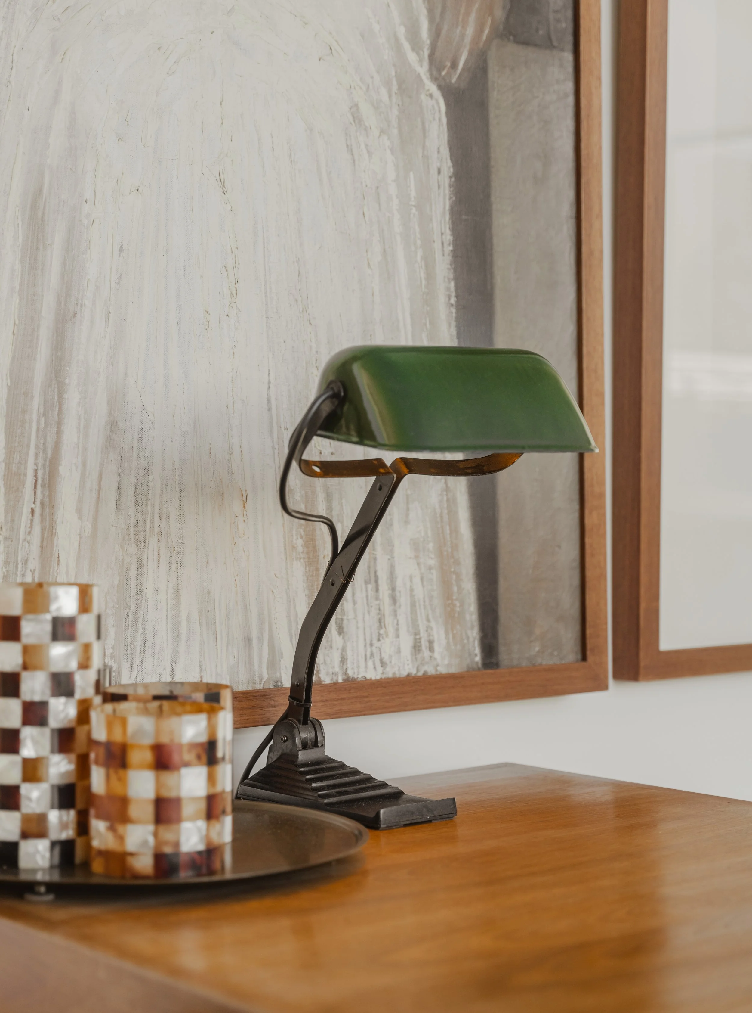 A vintage green desk lamp on a wooden surface next to colorful mosaic candles and a framed abstract painting in the background.