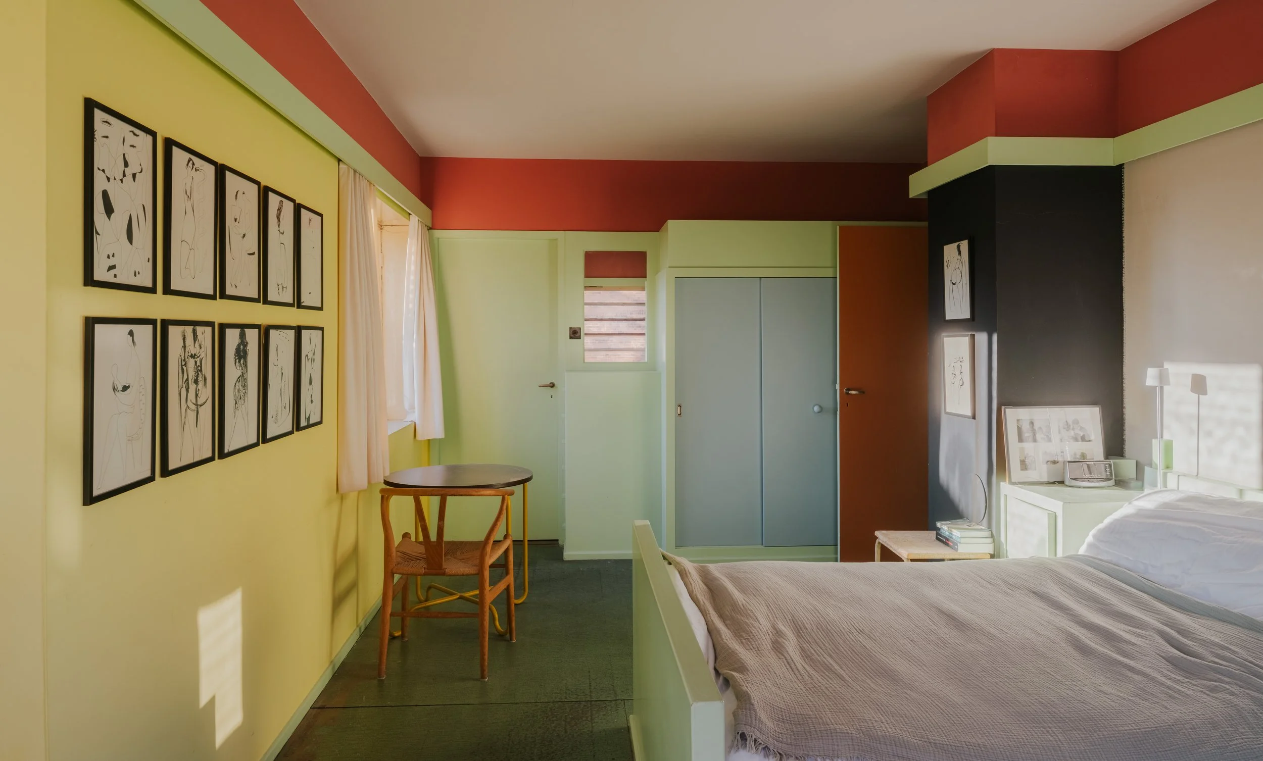 A bedroom with colorful walls, framed abstract artwork, a bed with beige blanket, a nightstand with books and a telephone, a small round table, and a wooden chair, with a window and closet.