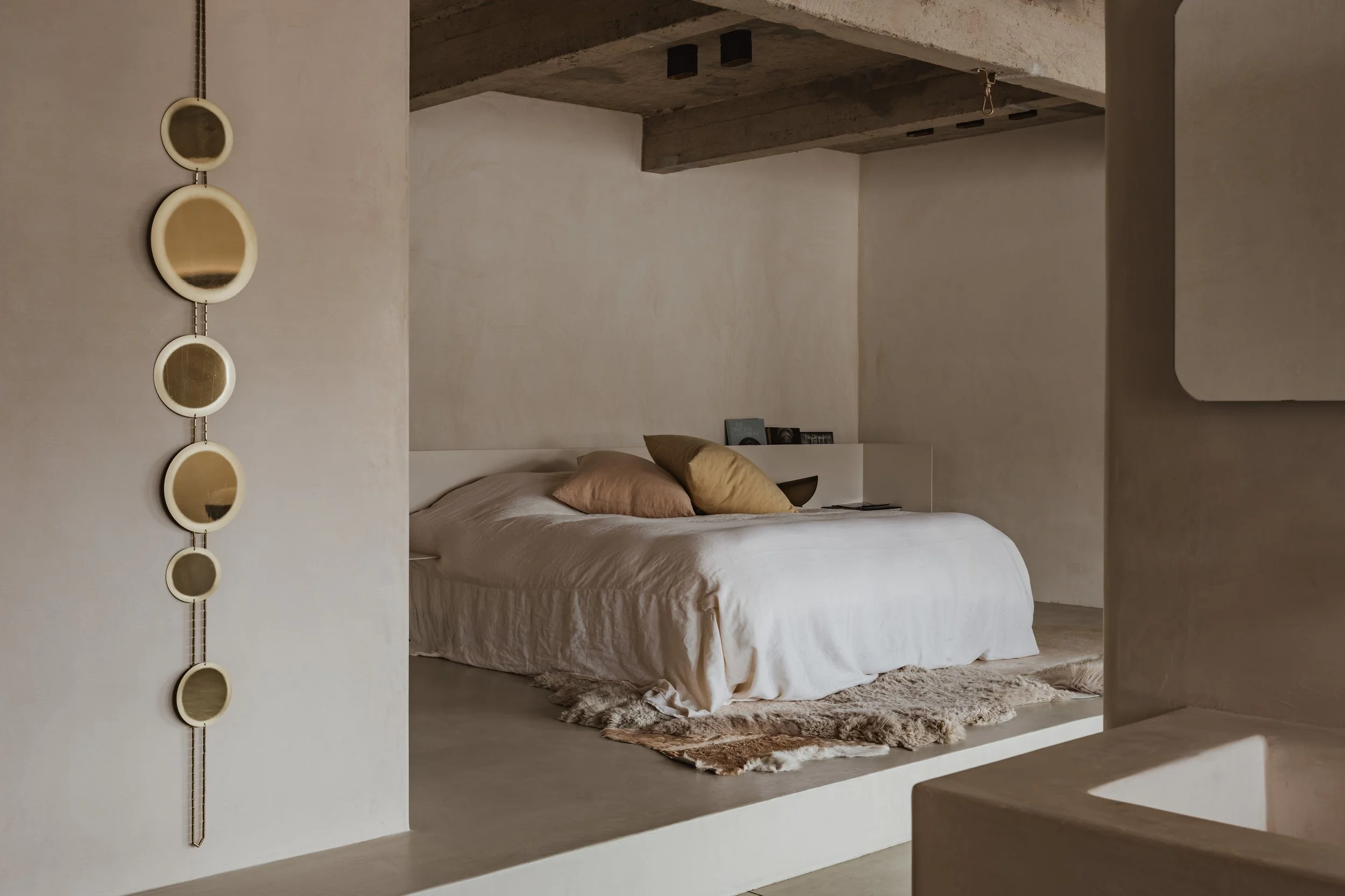 A minimalist bedroom with a white bed, beige pillows, and a natural fiber rug, framed by textured, cream-colored walls and a concrete ceiling.