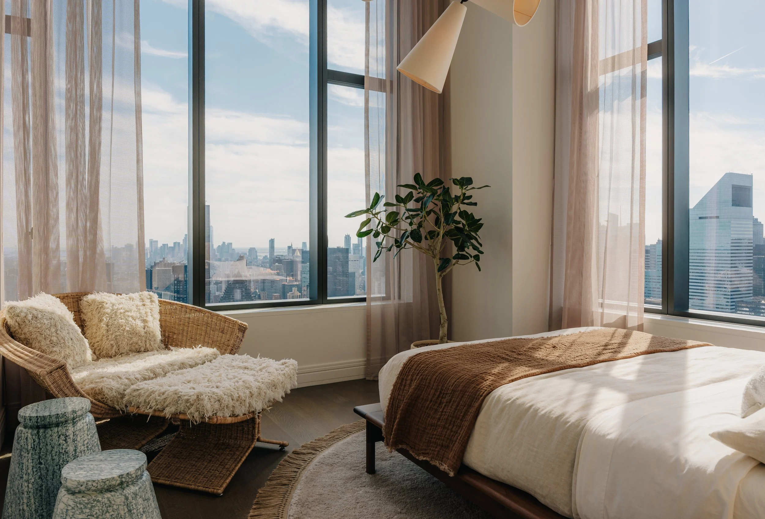 A bedroom with large windows showing a city skyline, a bed with a brown blanket, a wicker chair with fuzzy pillows, a green potted plant, and sheer curtains.