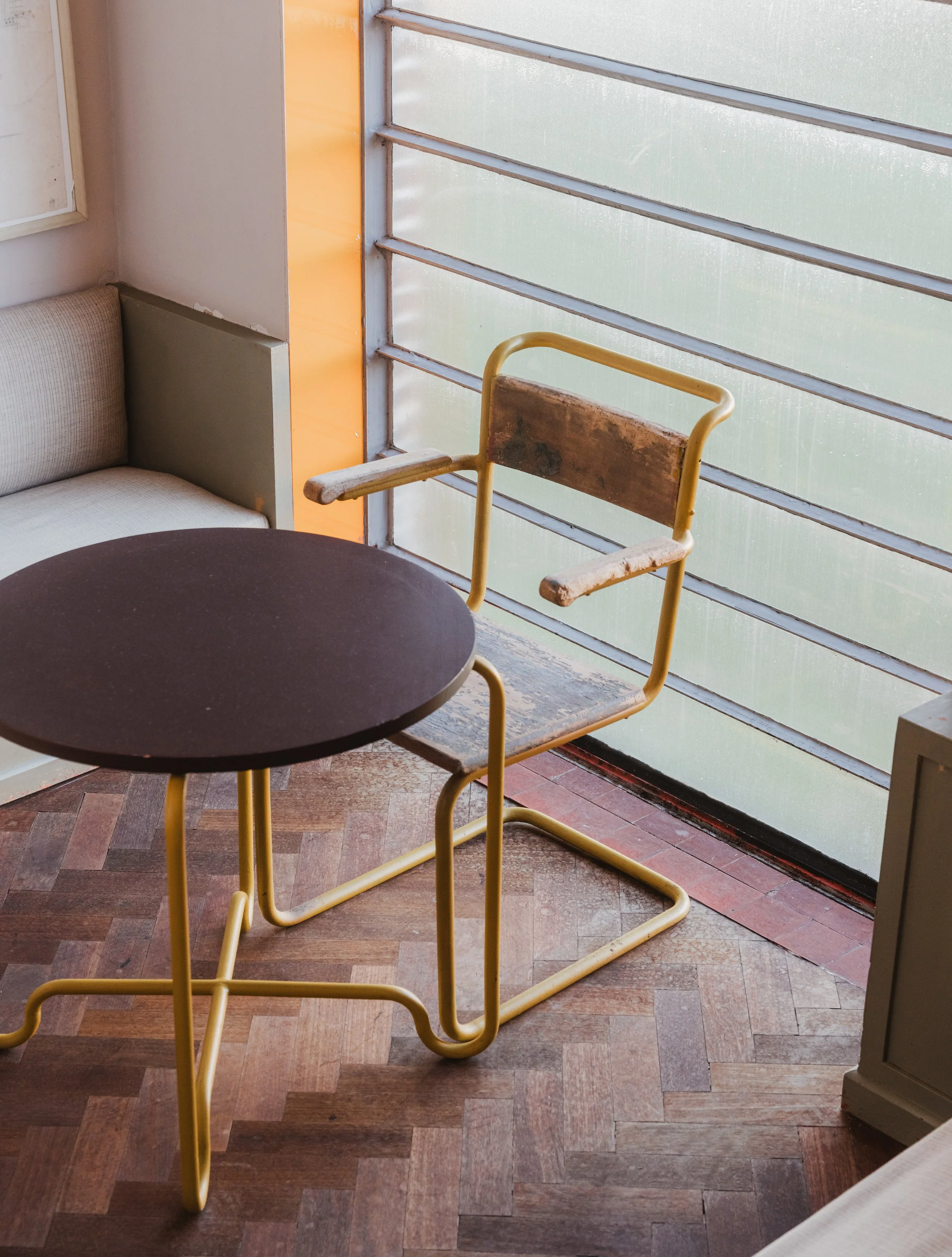 Empty yellow chair with wooden armrests and seat, next to a small black round table in a sunlit room with wood flooring and large window with horizontal metal slats.