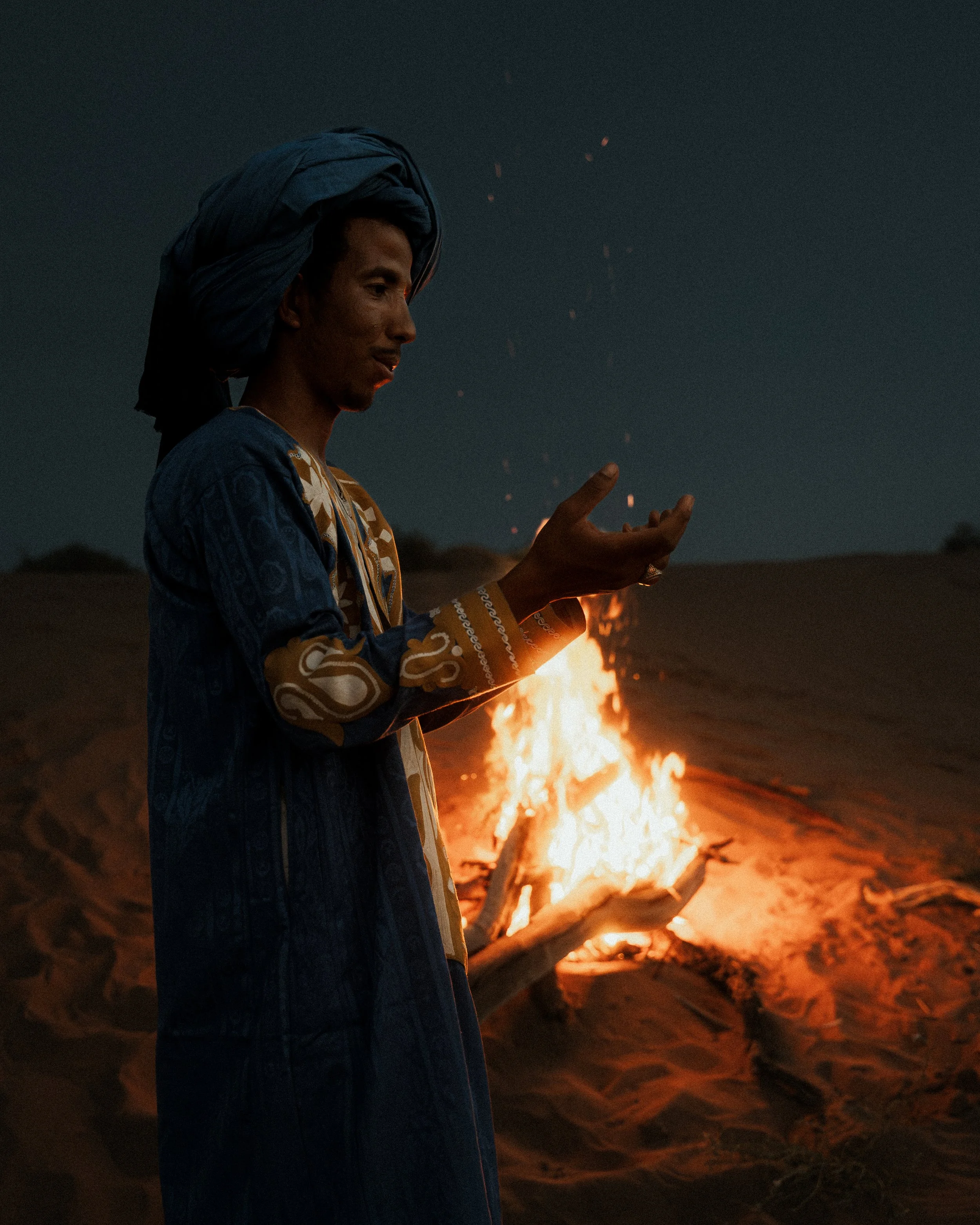 A person wearing traditional clothing and head wrap standing near a campfire at night, with sparks flying into the dark sky.