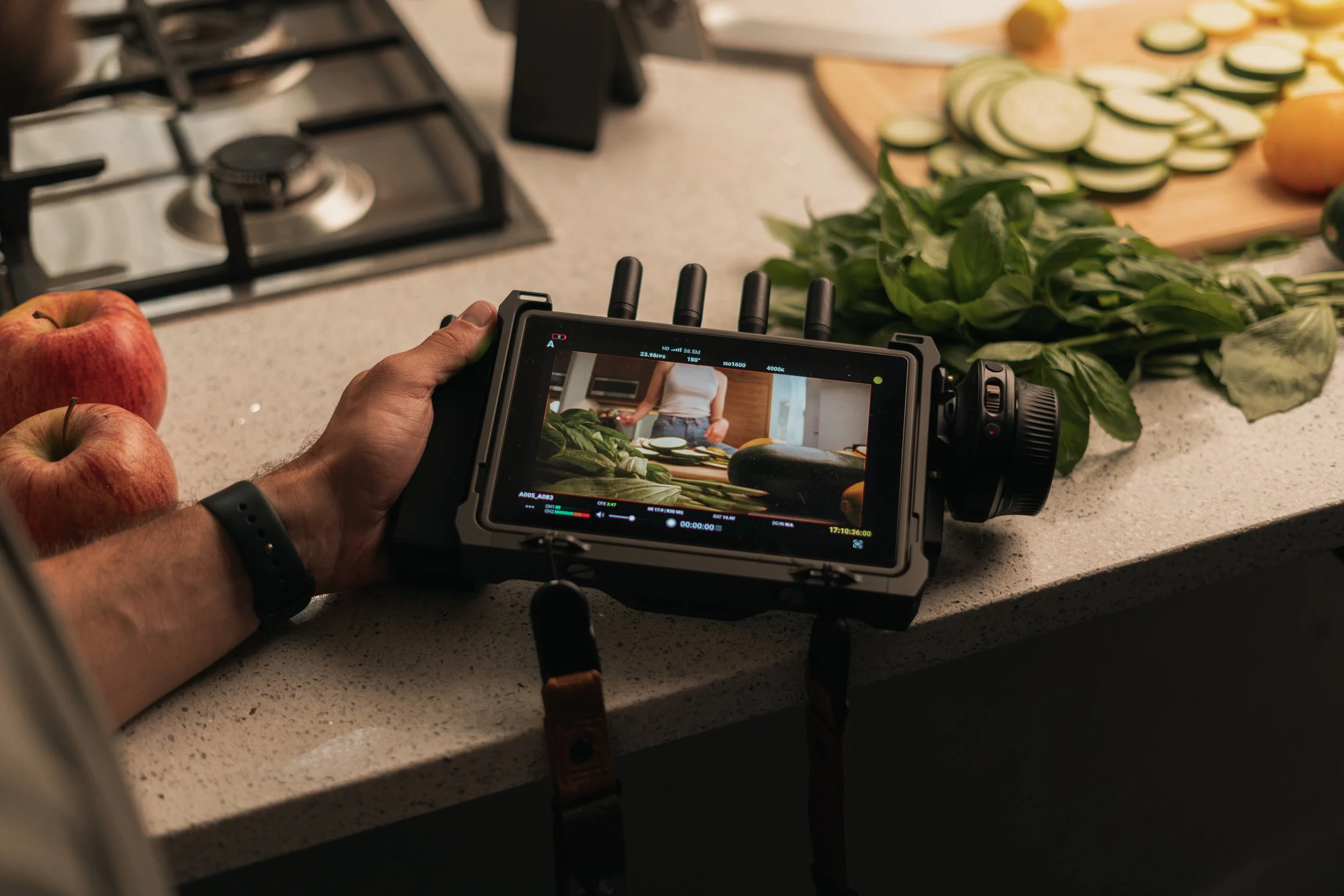 A person filming a cooking video in a kitchen, with fresh vegetables and apples on the countertop.