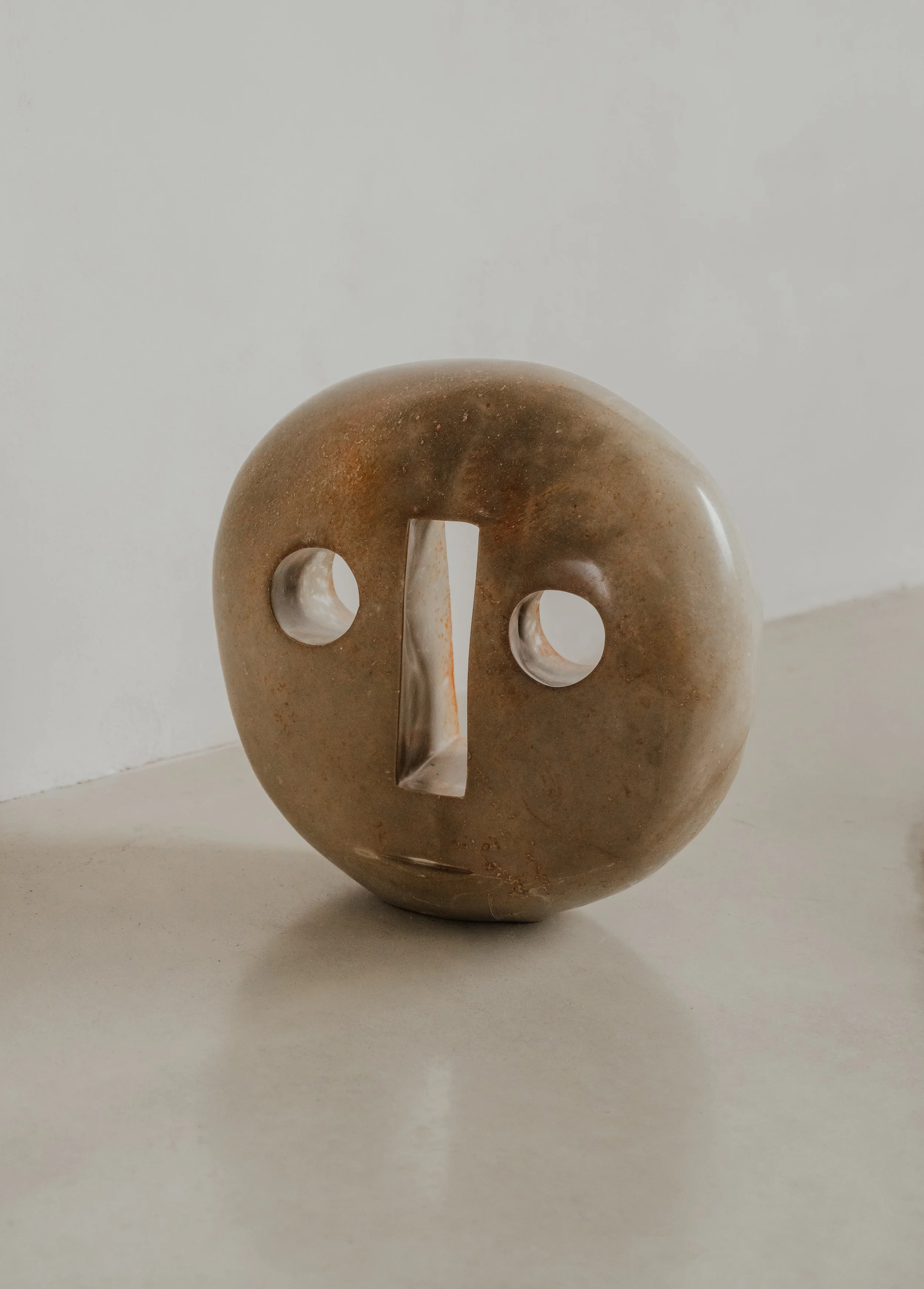 A rounded stone sculpture with two circular cutouts and a rectangular opening in the center, resembling a face, placed on a white surface against a plain white wall.