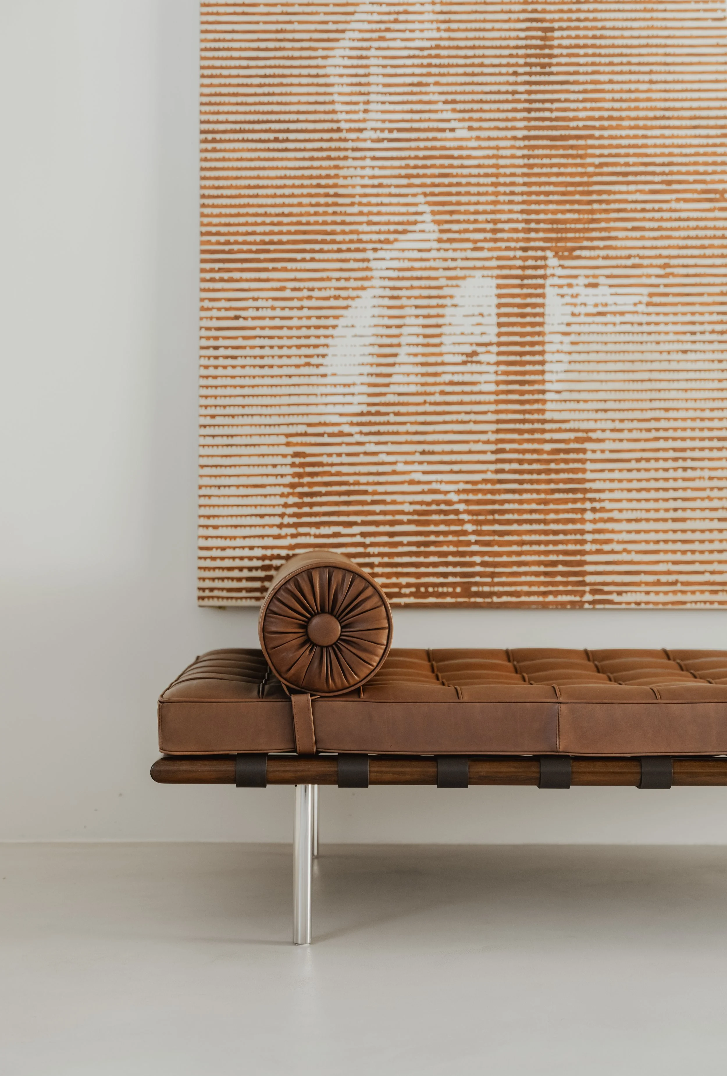Brown leather tufted bench with a round leather pillow and a framed portrait artwork in the background.