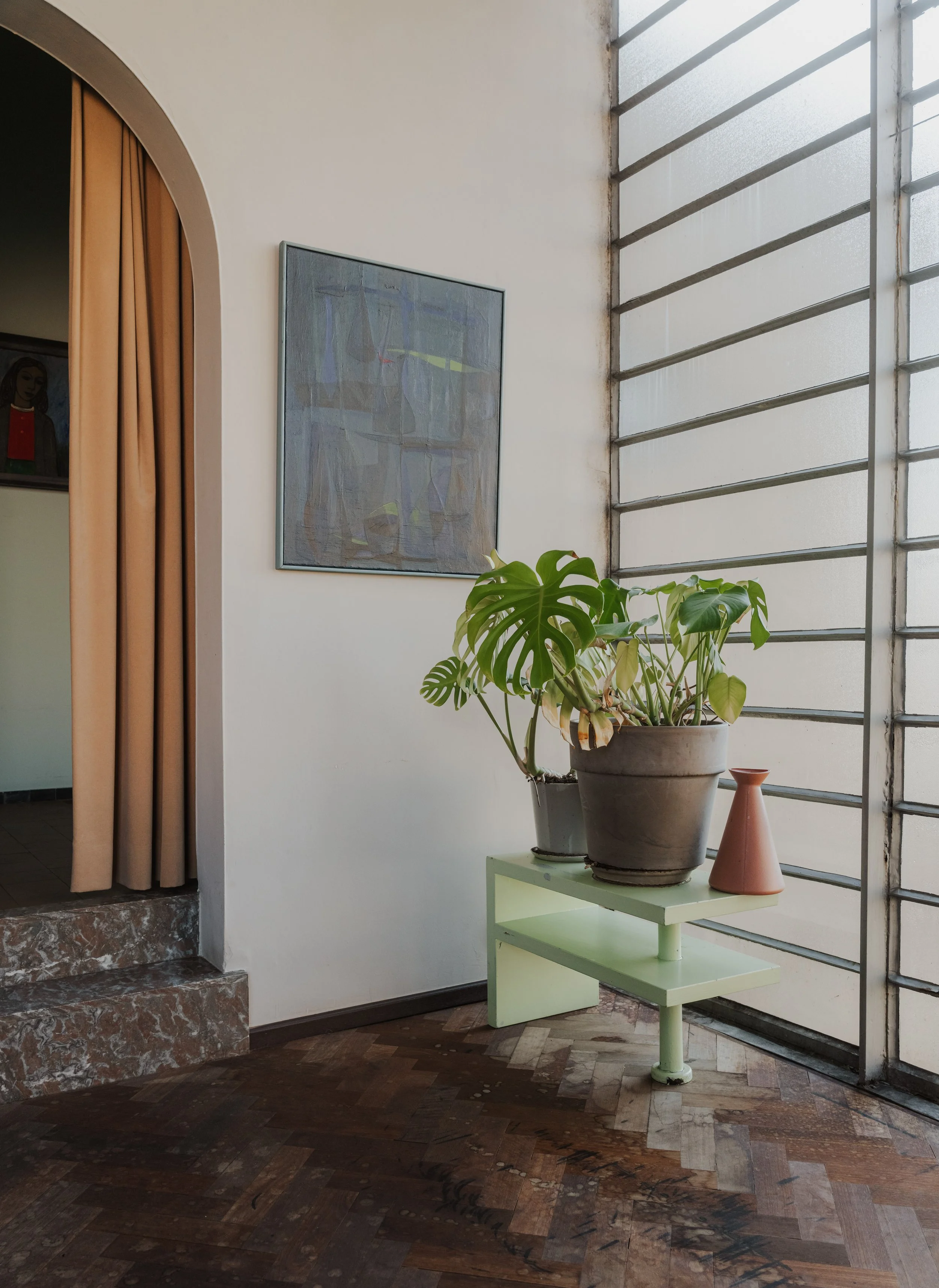 A corner of a room with a light green bench holding two potted plants and a small terra cotta vase, next to a large window with horizontal metal bars, and a framed abstract painting on the wall.
