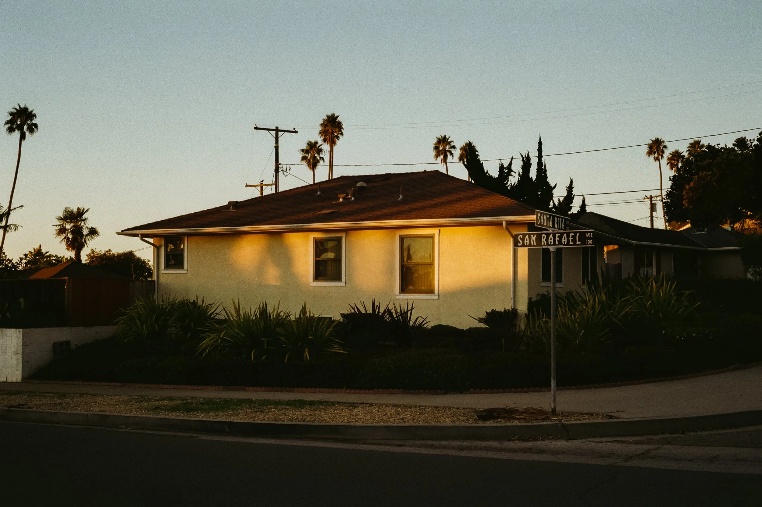 A single-story house on a corner lot at the intersection of Santa Rita and San Rafael streets during sunset, with palm trees in the background and a garden with plants in front.