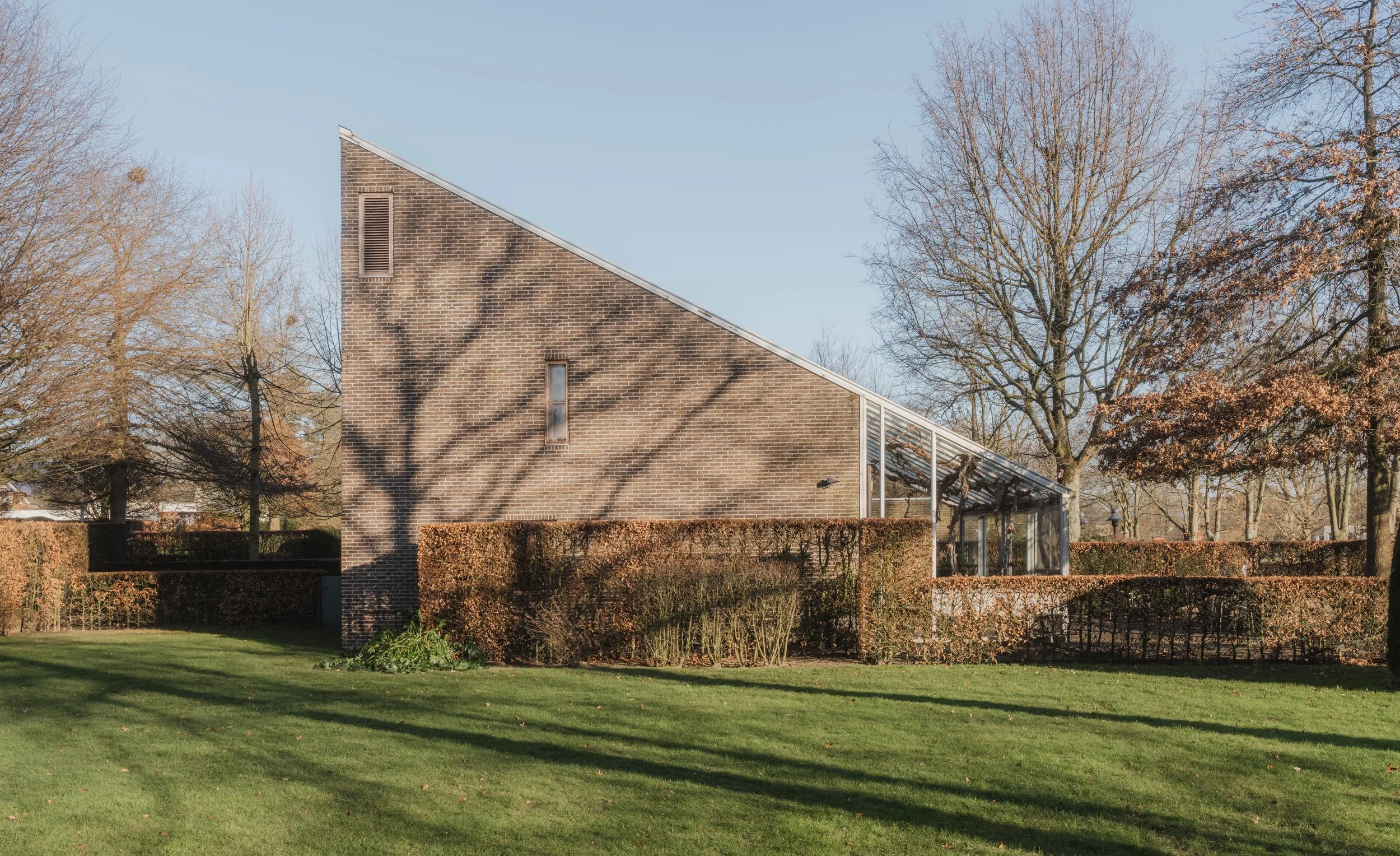 Side view of a modern house with a sloped roof, brick walls, and a glass-enclosed sunroom, surrounded by trees and hedges, on a clear day with shadows on the lawn.