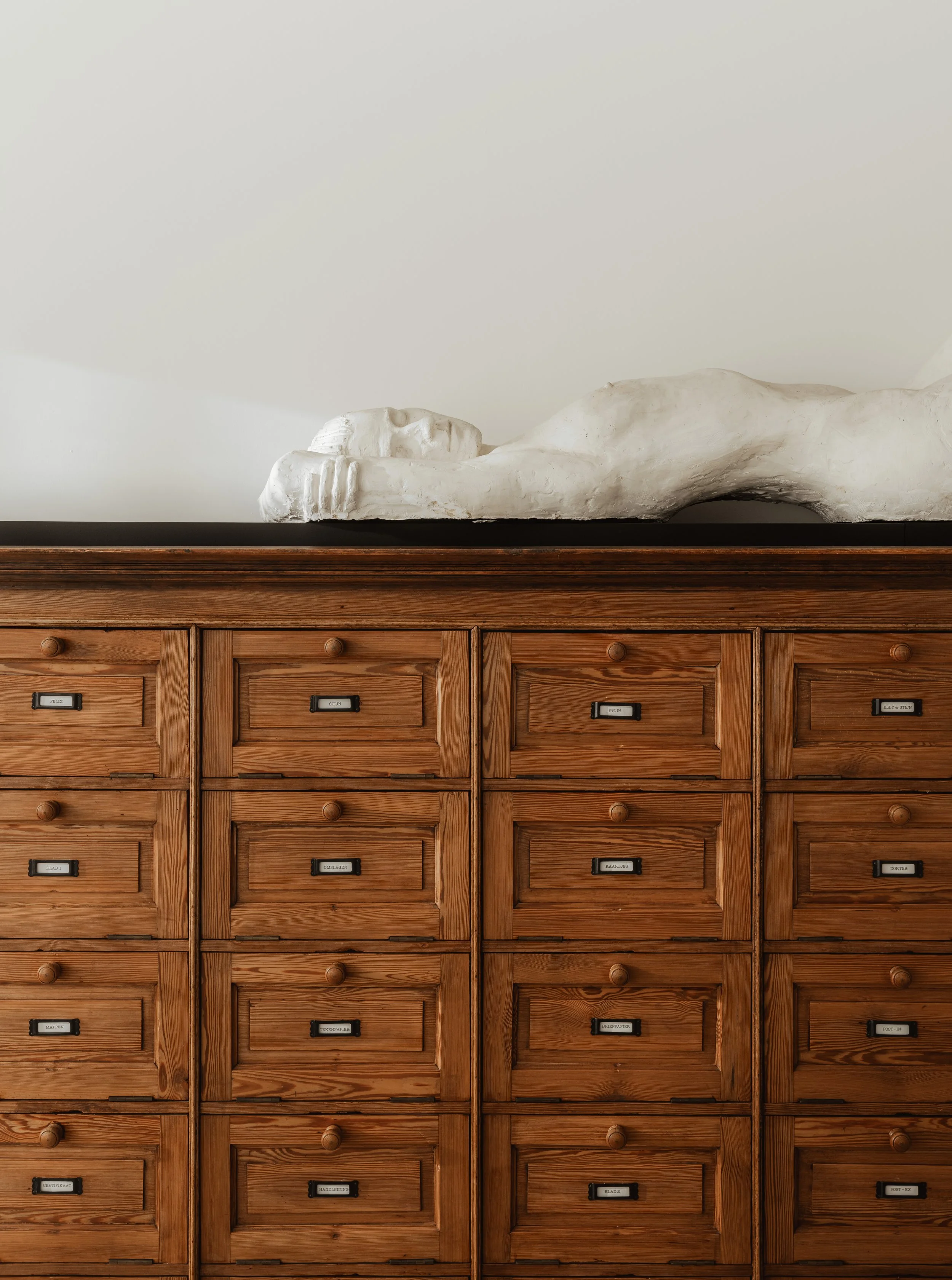 A wooden cabinet with multiple small drawers, labels on each drawer, and a reclining white sculpture of a human figure on top.