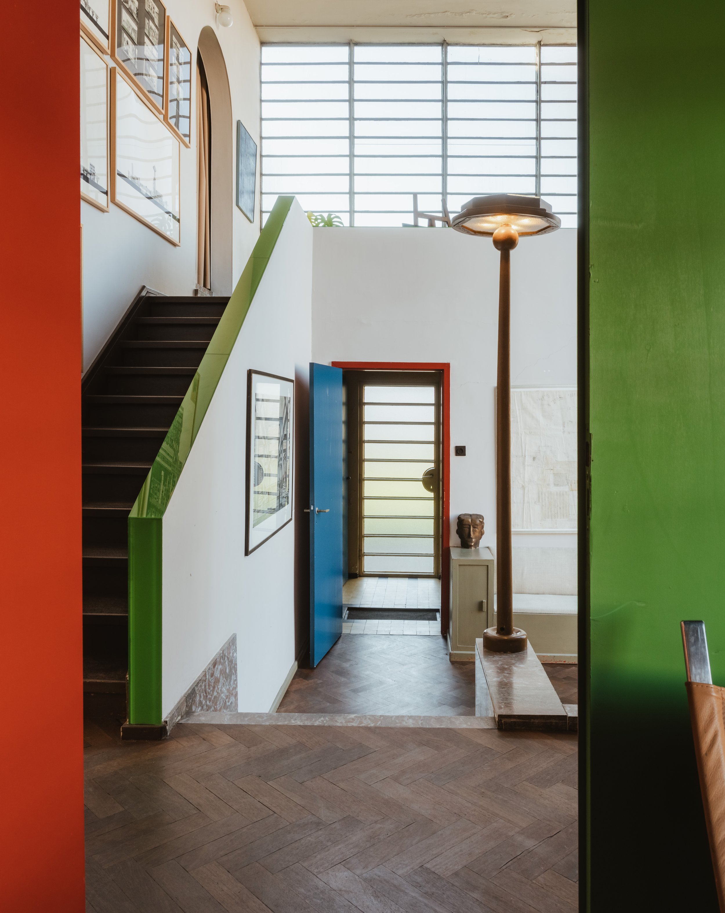 Interior view of a modern, colorful home with a staircase on the left, artwork on the wall, and a brightly lit entrance door at the back. Green, orange, and blue accents add a vibrant touch.