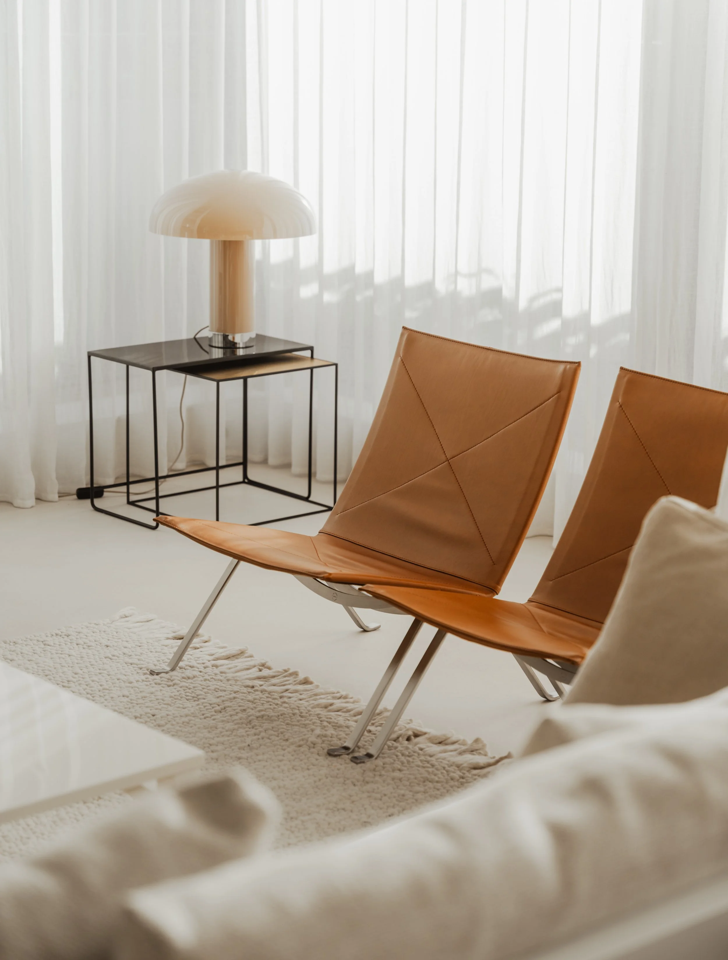 Living room interior with beige leather chairs, side table, modern lamp, white curtains, and cream-colored rug.