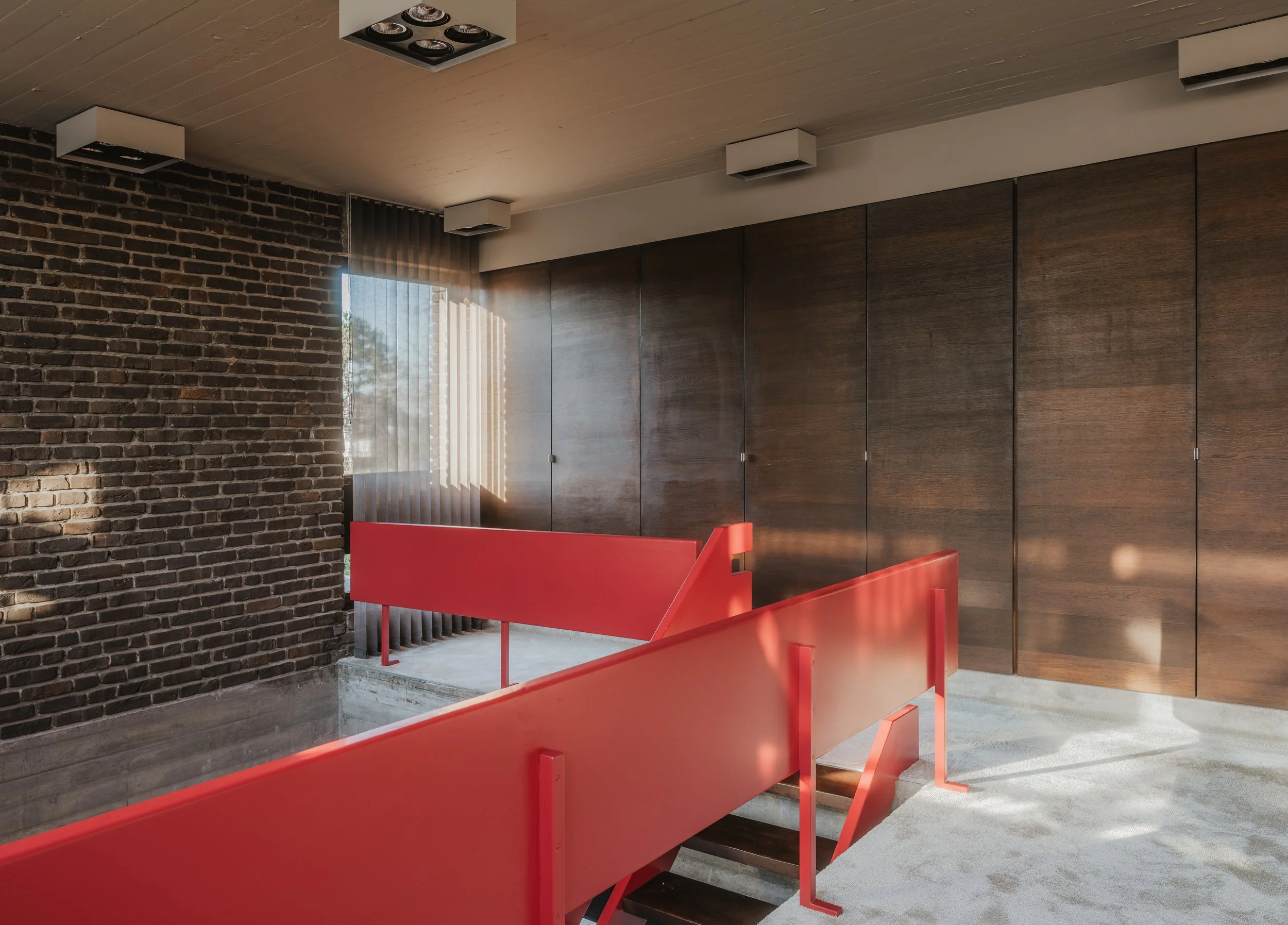 Indoor staircase with red railings, wooden wall panels, a brick wall on the left, and a window with vertical blinds letting in sunlight.