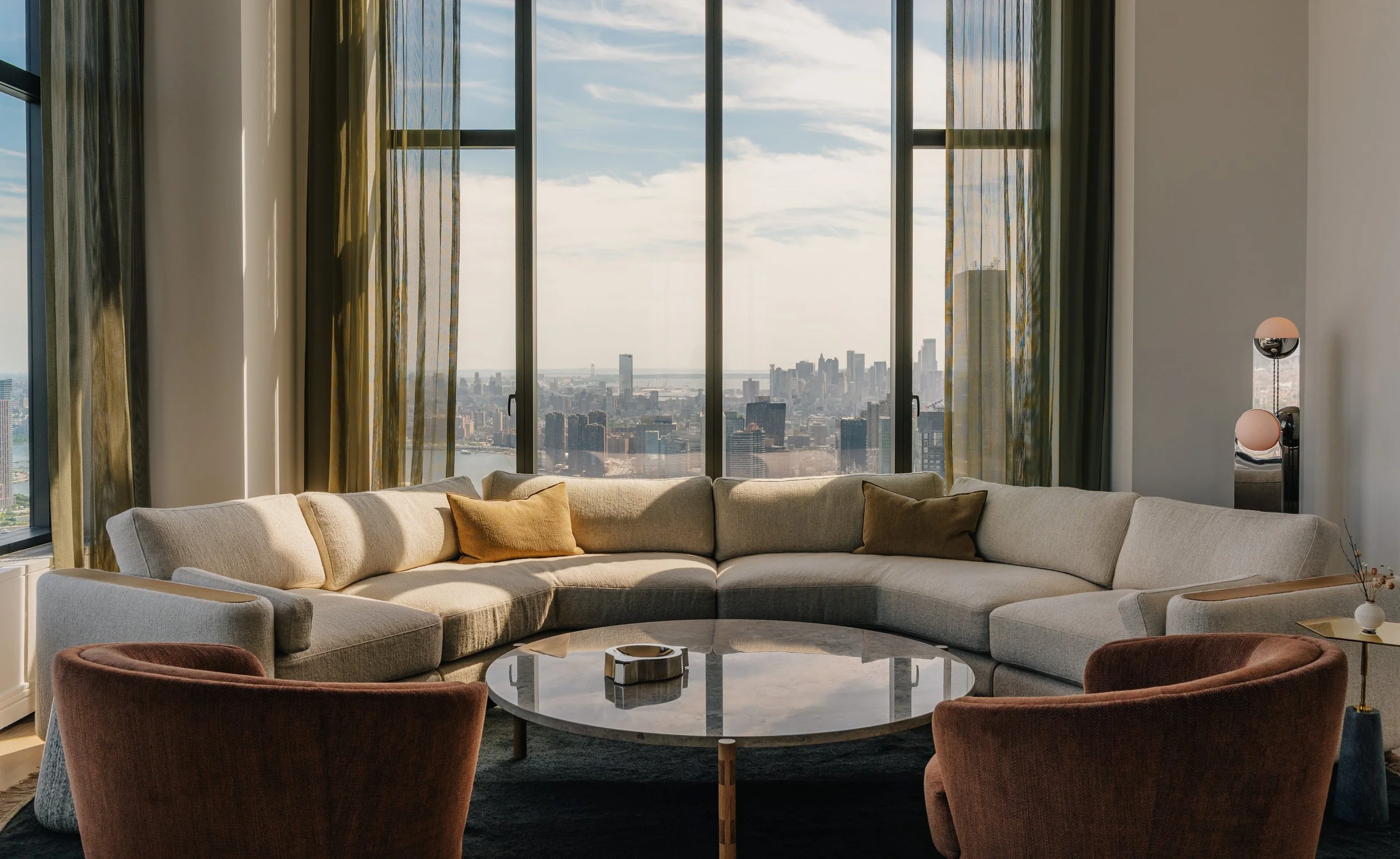 Modern living room with large windows showing city skyline and water view, beige sectional sofa with throw pillows, round marble coffee table, and velvet armchairs.