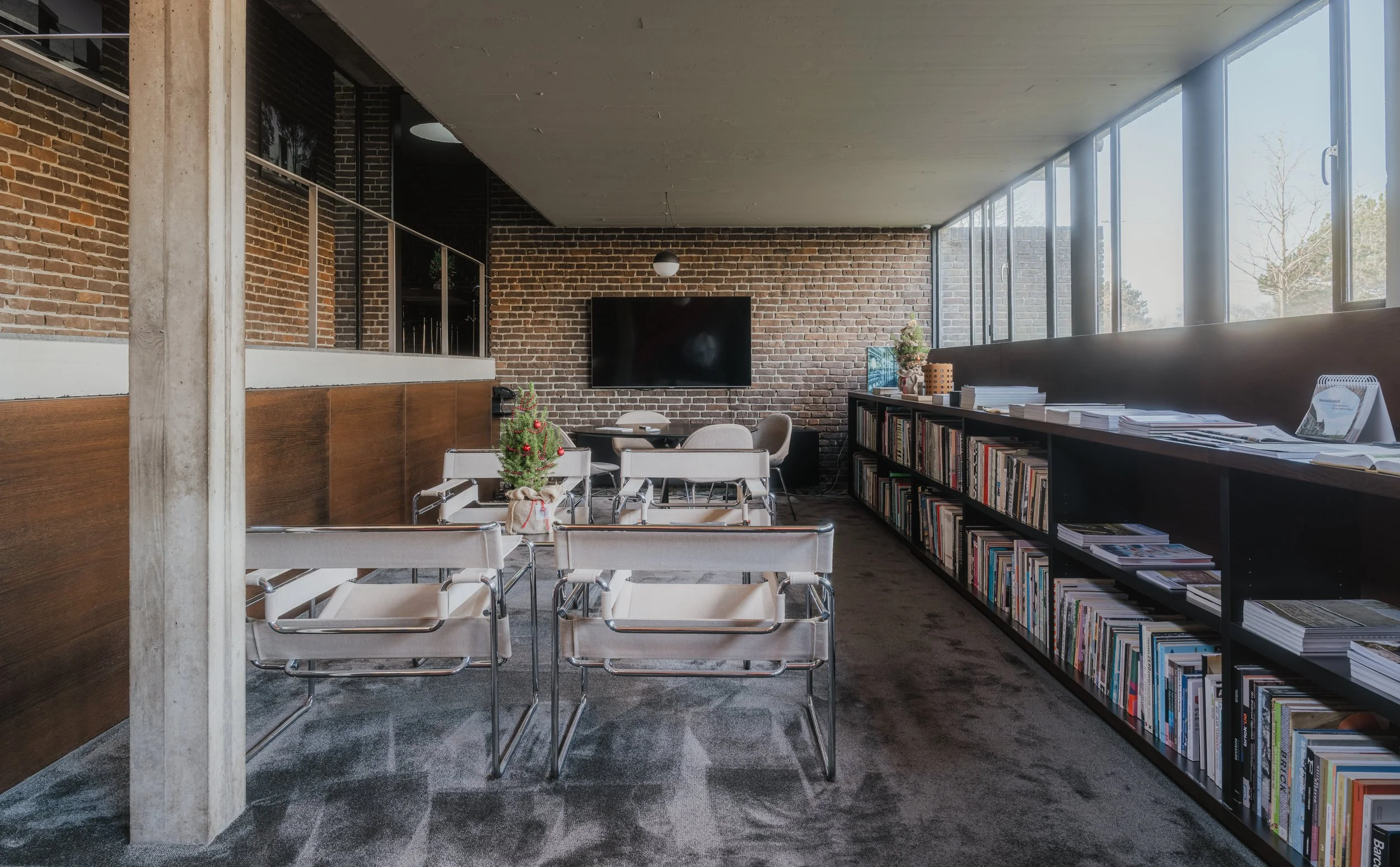 A modern meeting room with brick walls, a large window, and a bookshelf filled with books. There are chairs arranged around a small table with Christmas decorations.