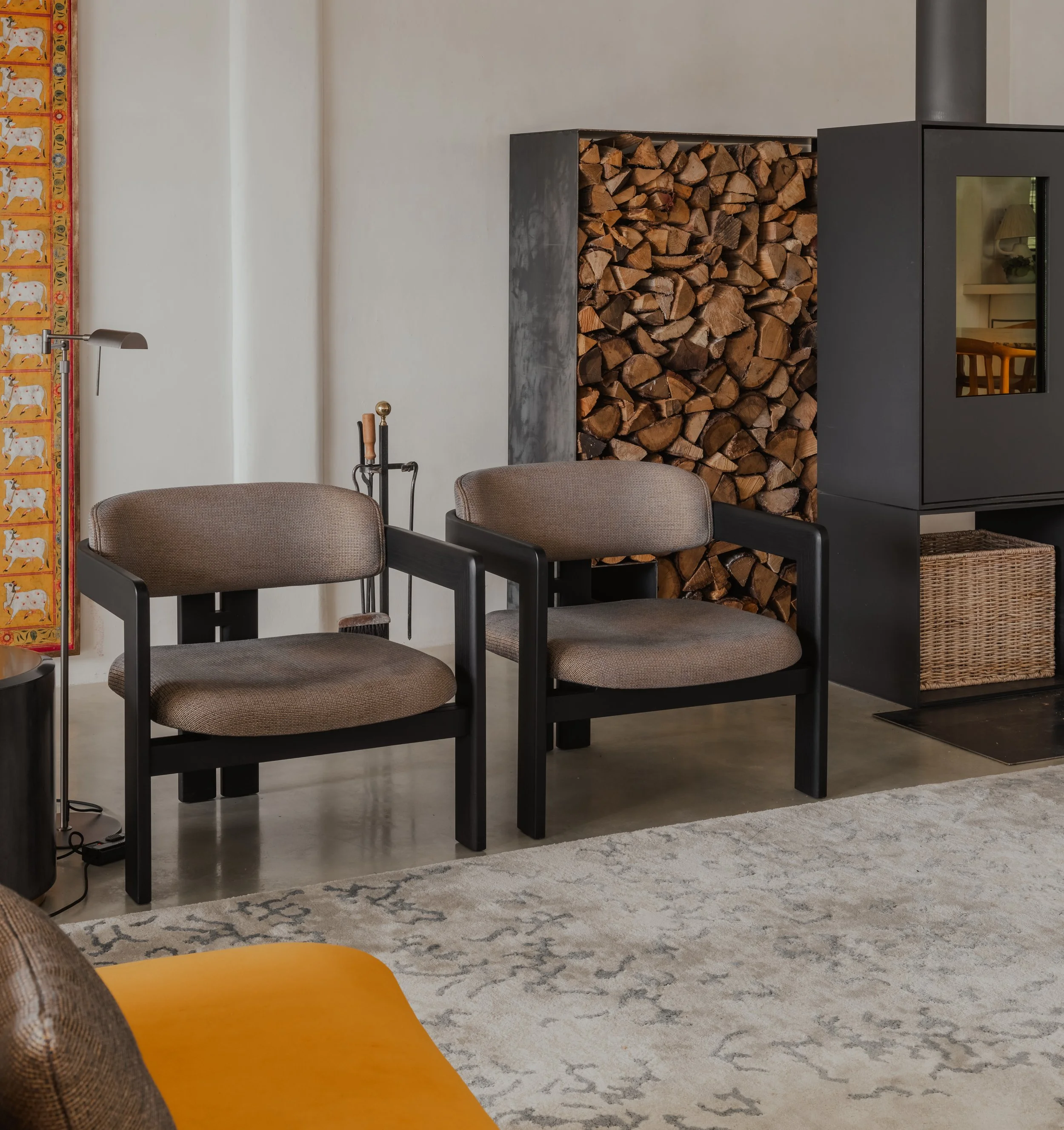 Living room with two beige upholstered armchairs, a wood-burning stove with firewood, a patterned rug, a yellow couch, and a decorative horse-themed wall hanging.