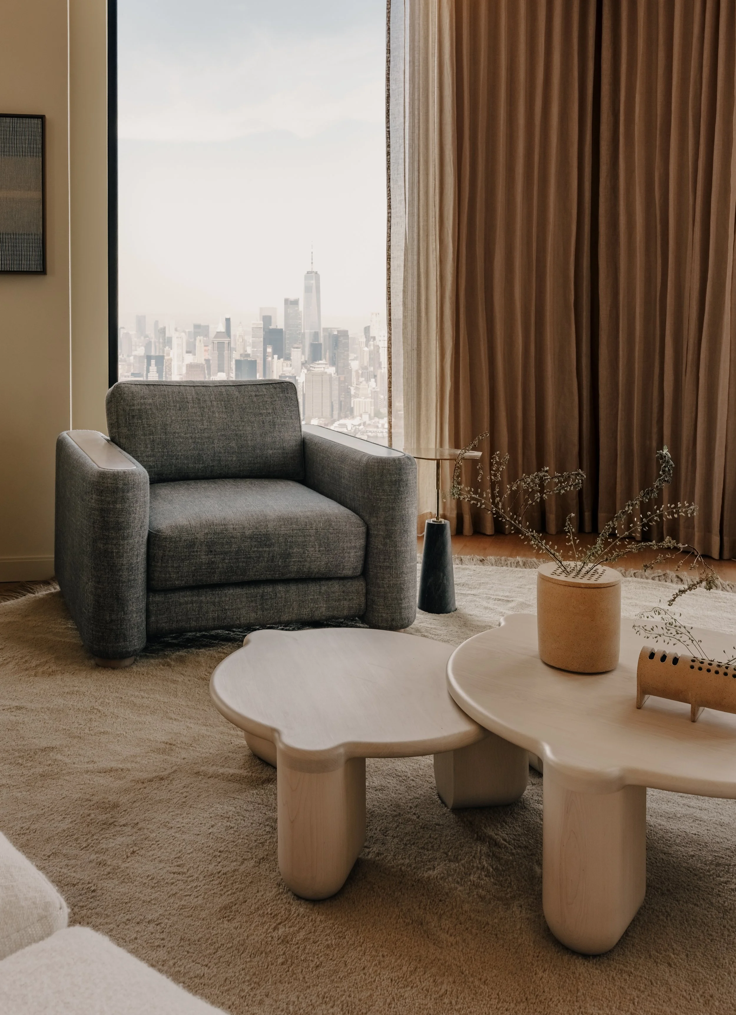 Living room with a gray armchair, two white coffee tables, potted plant, beige carpet, large window with city skyline view, beige curtains.