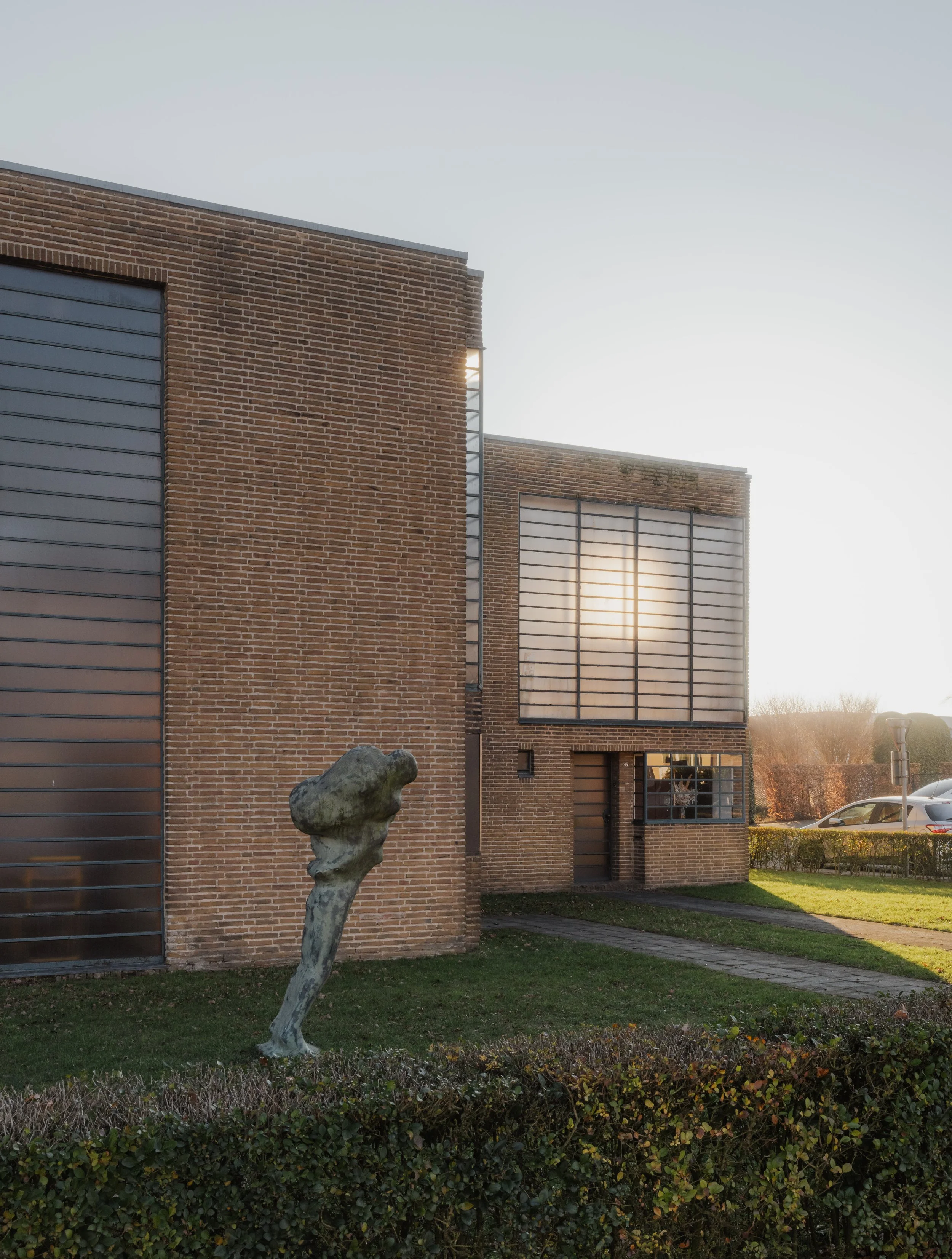 A modern brick building with large windows and a sculpture of an abstract shape in front. The sun is rising or setting in the background.