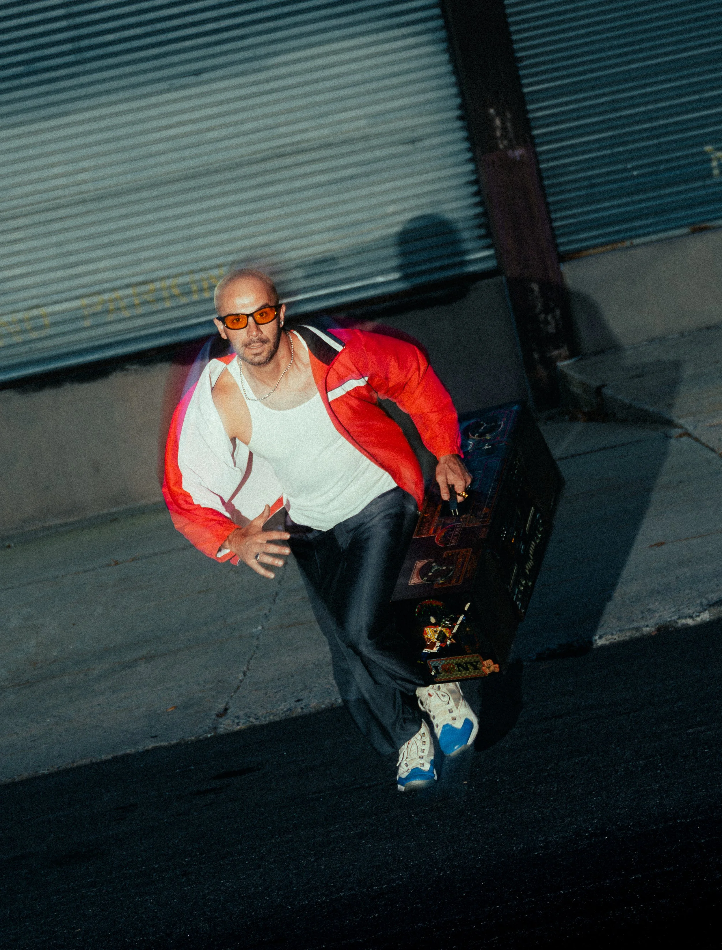 Man with blond hair wearing orange sunglasses, a white tank top, red jacket, black pants, and sneakers, standing on a sidewalk next to a sound system, with his hand on his hip.