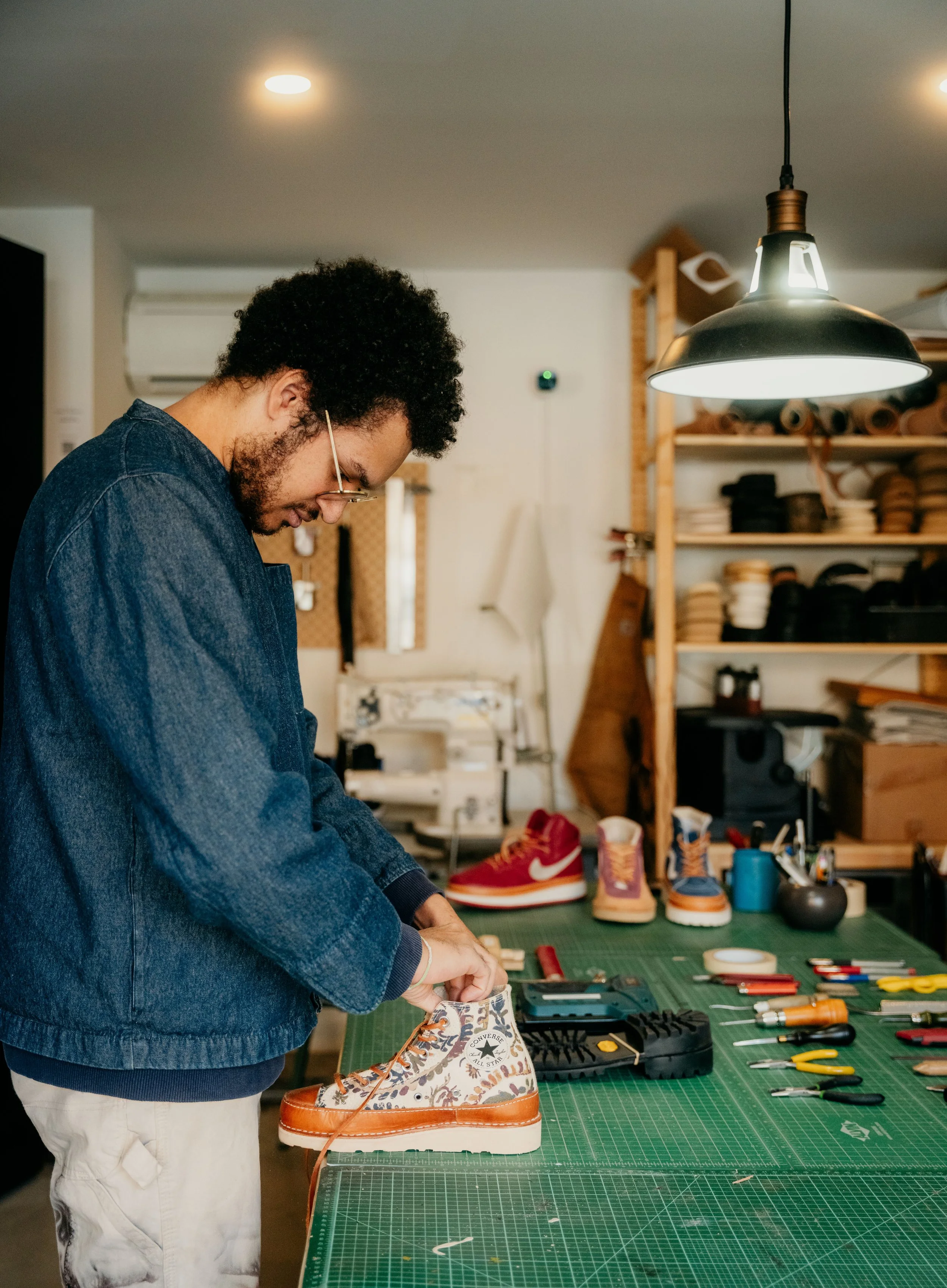 A man working on customizing a pair of floral patterned sneakers in a workshop with tools and shoes on the table and shelves.
