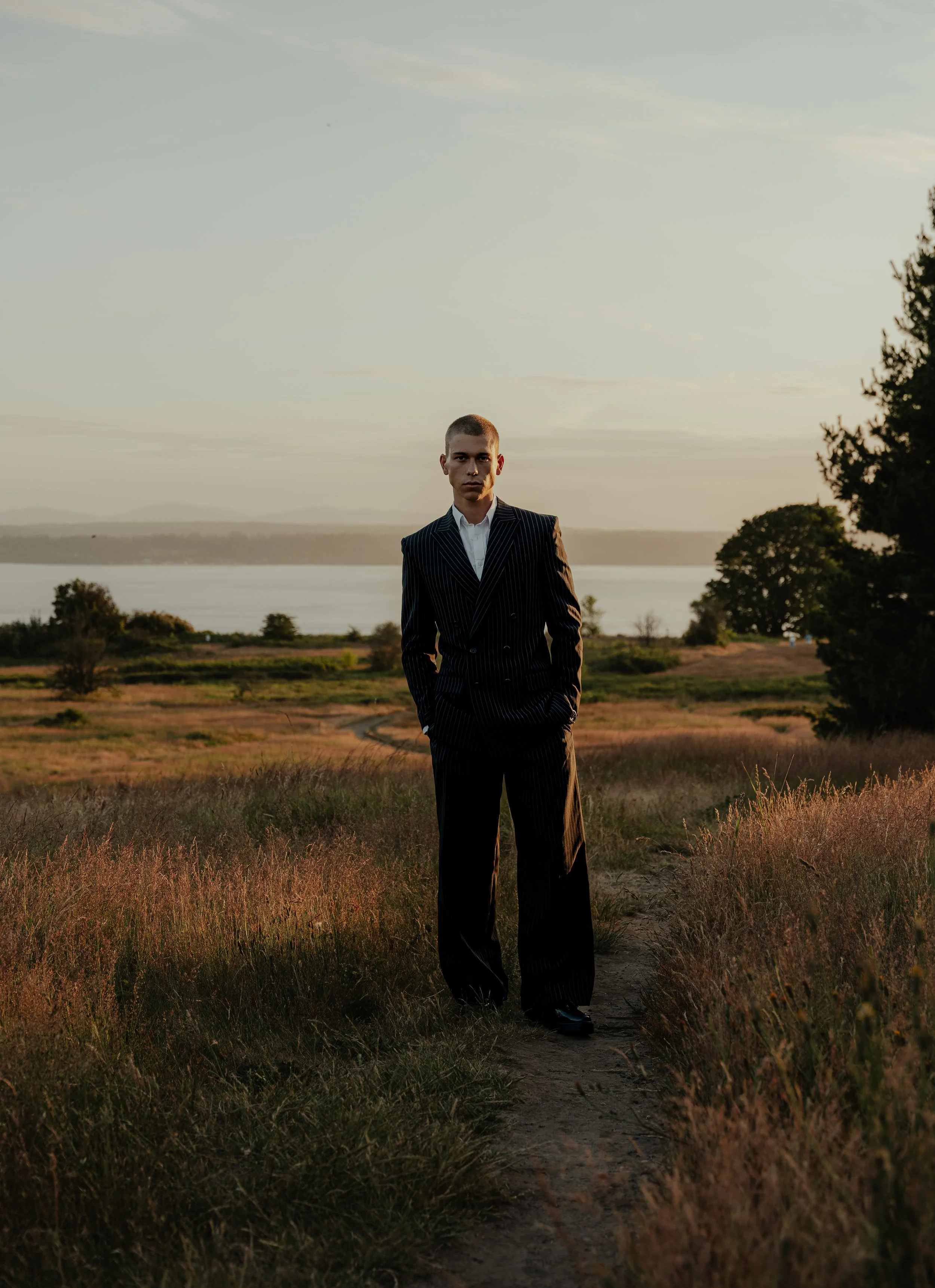 A young man in a pinstripe suit standing on a dirt path in a grassy field at sunset with trees and a body of water in the background.