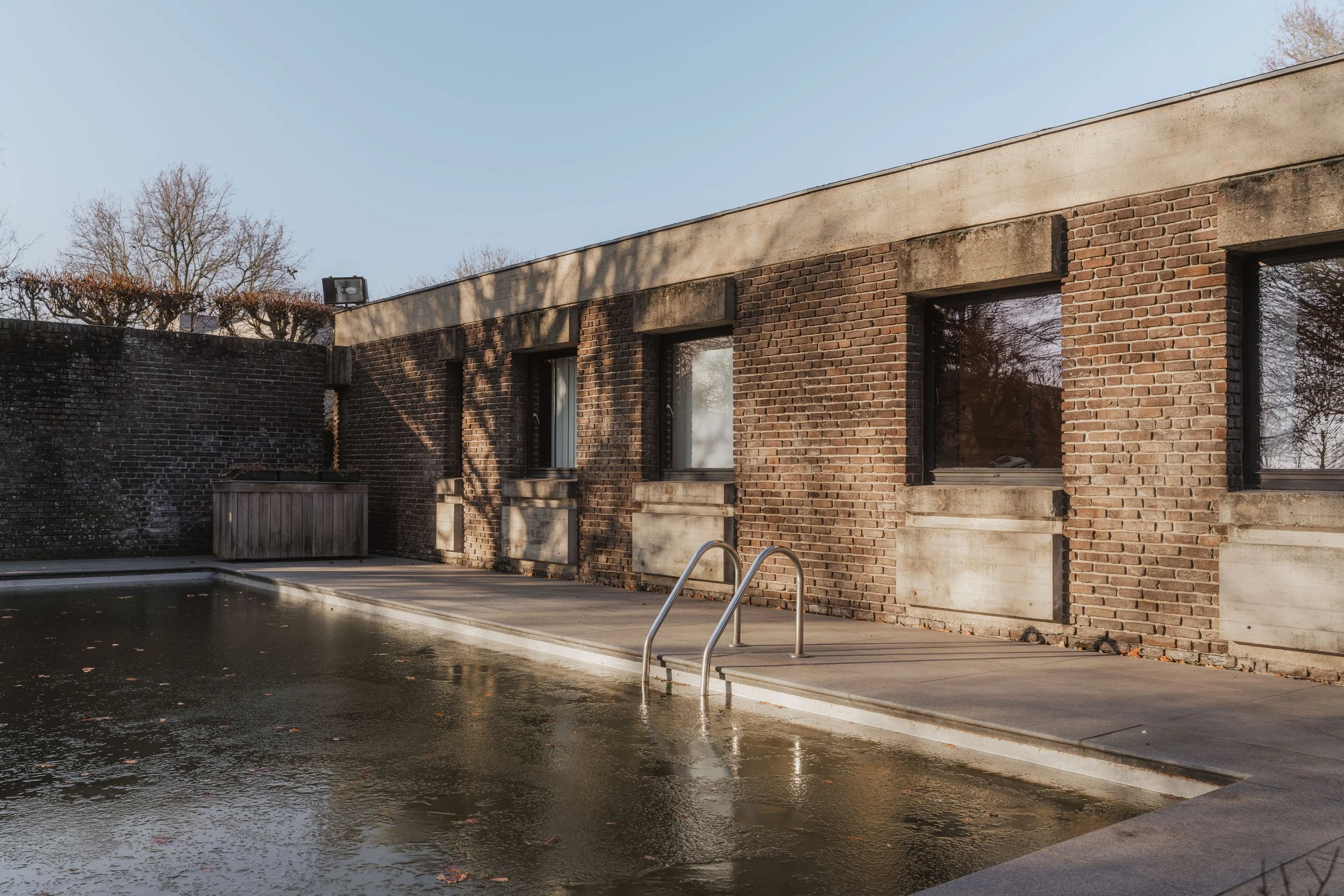 Exterior of a brick building with large windows next to a swimming pool. The scene appears to be during late fall or winter, with bare trees and a clear sky.
