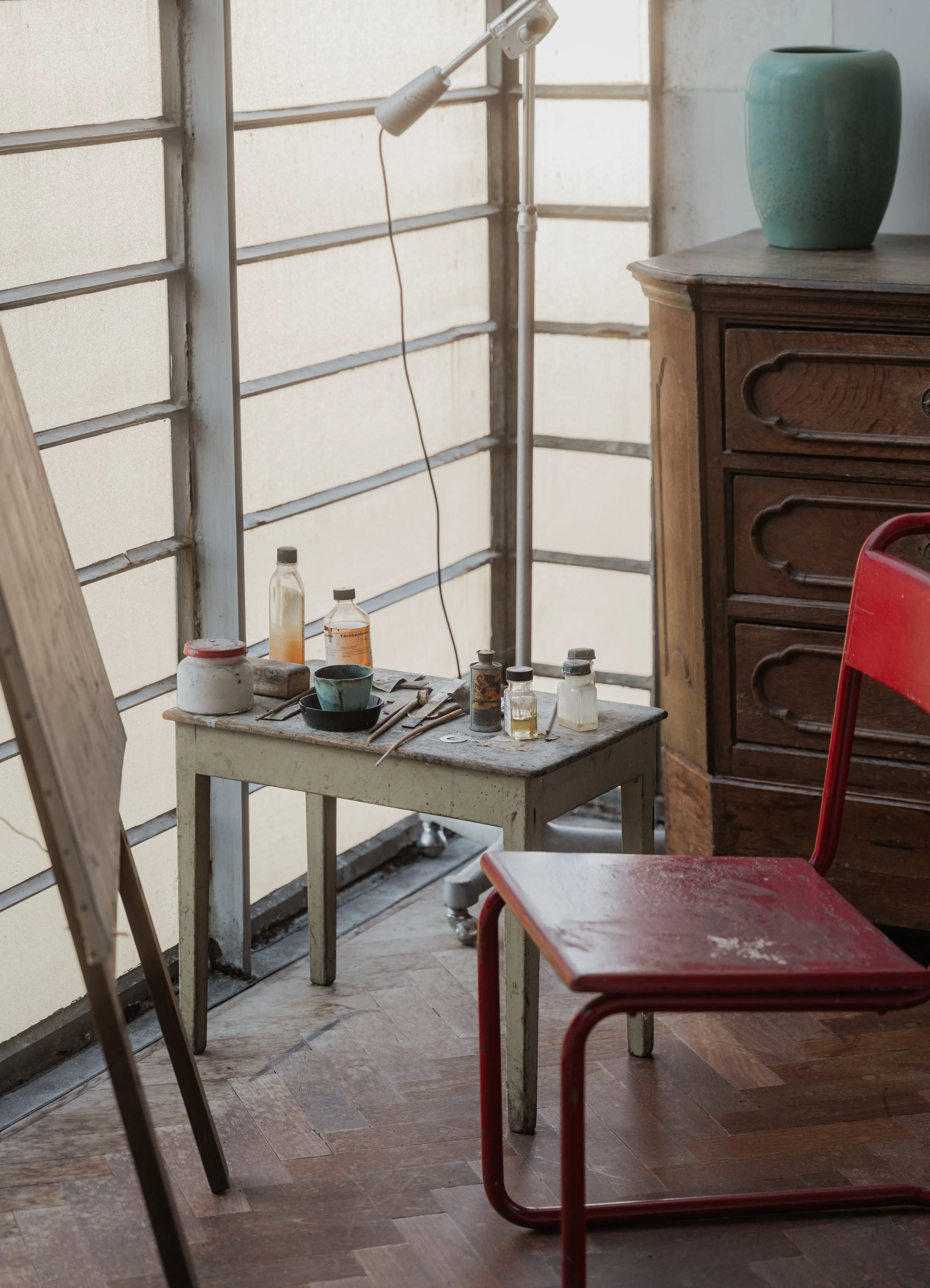 A small, old wooden table with jars, bottles, and tools on top, next to a red metal chair and a wooden dresser, in a sunlit room with frosted glass window panels.