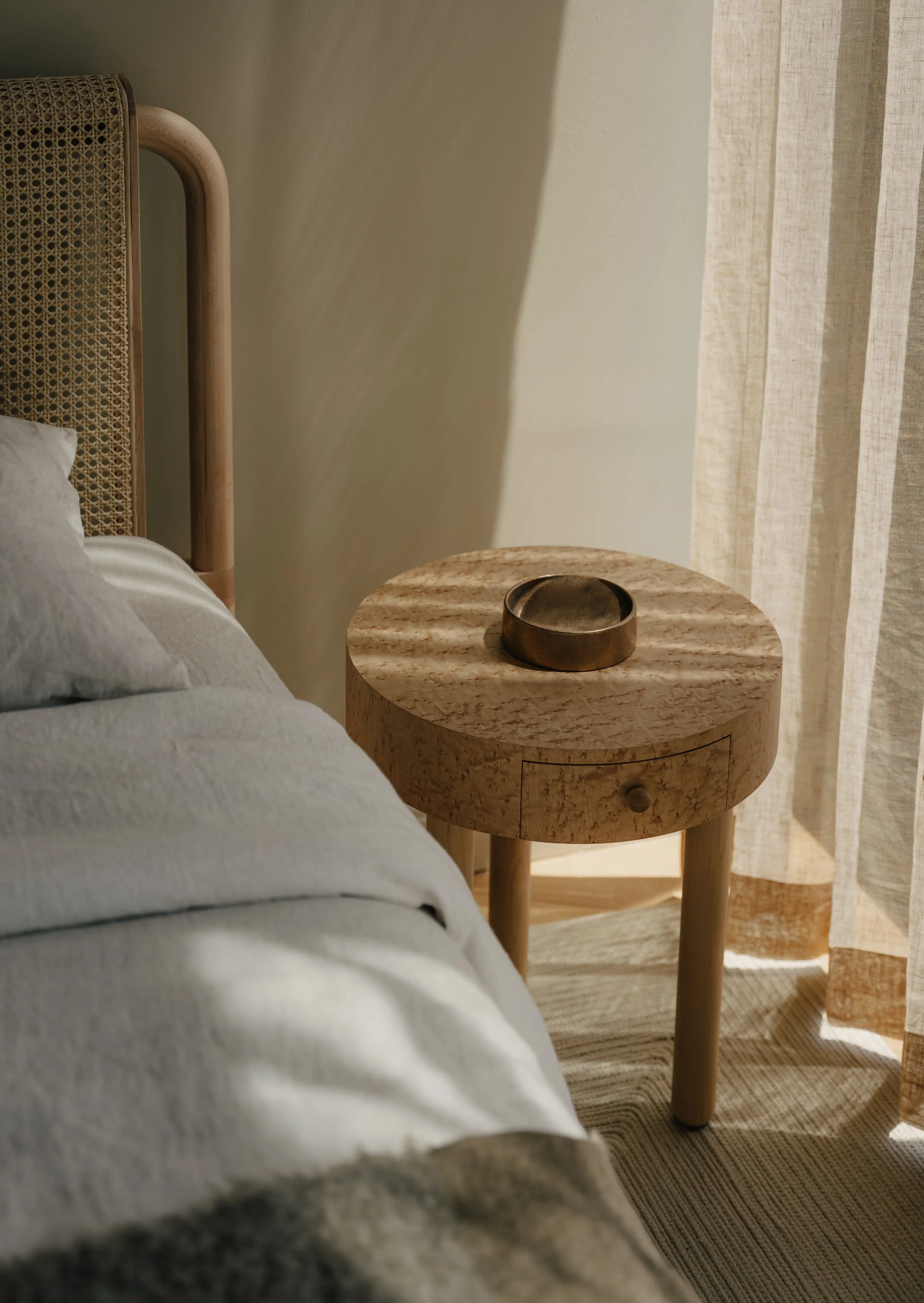 Close-up of a bedroom corner showing a bed with white linens, a round wooden bedside table with a metallic tray, a rattan headboard, and a beige curtain with vertical stripes.
