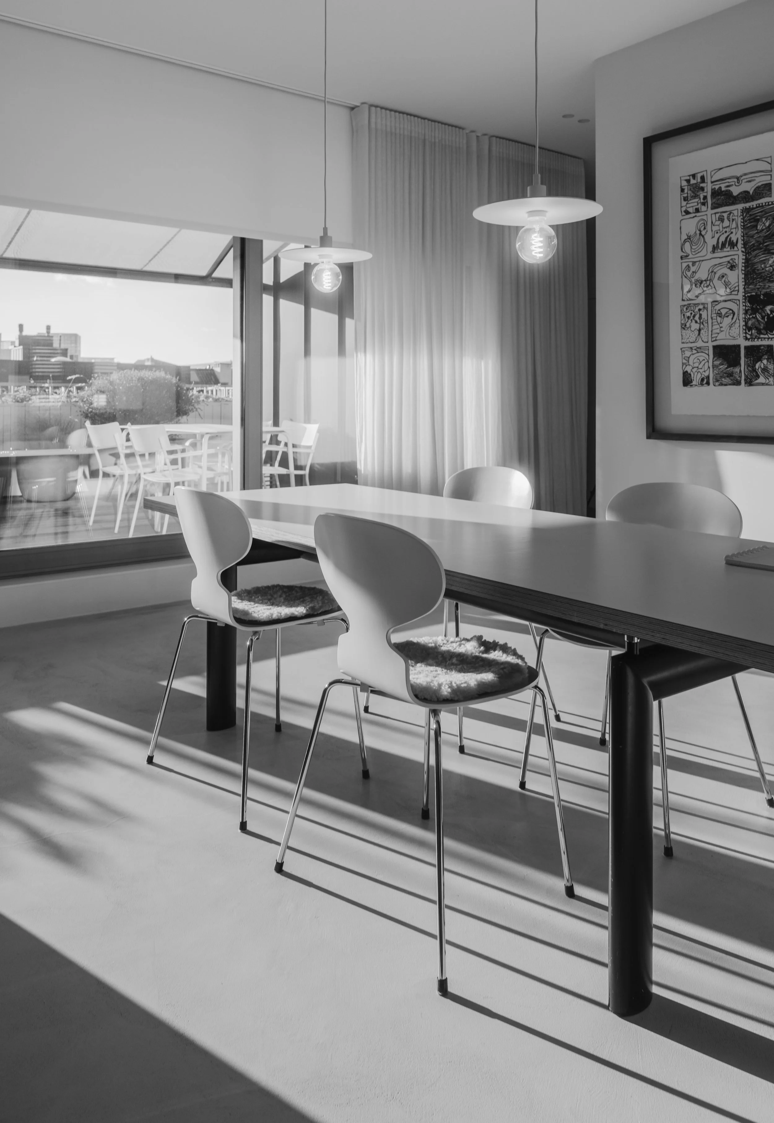Modern dining room with a table, chairs, and pendant lighting. Large sliding glass door leading to a balcony with outdoor seating and city view, framed with curtains. Black and white photo.