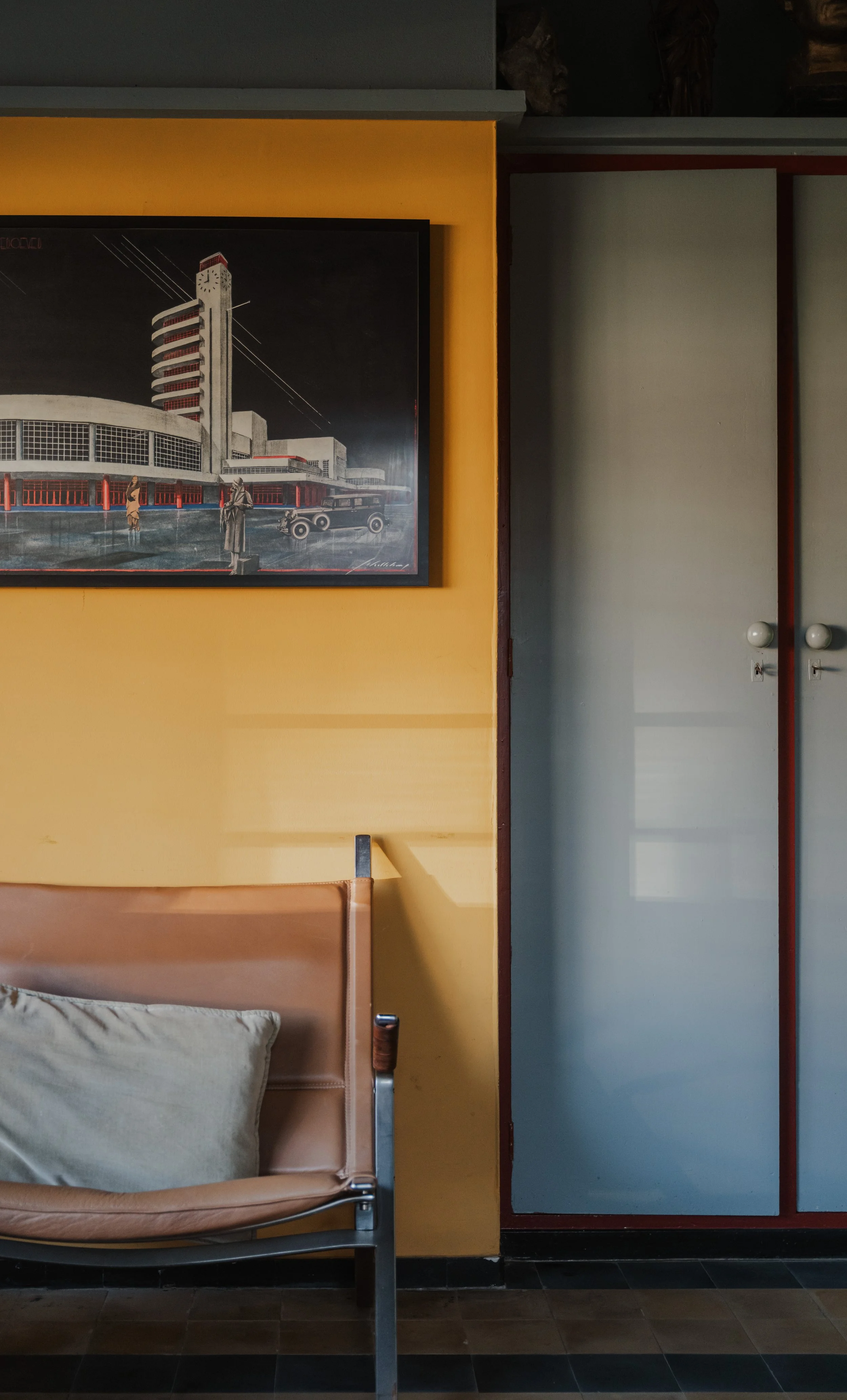Interior of a room with a beige leather sofa, a yellow wall with a vintage-style framed picture of a building, and a closed light blue closet with white knobs.