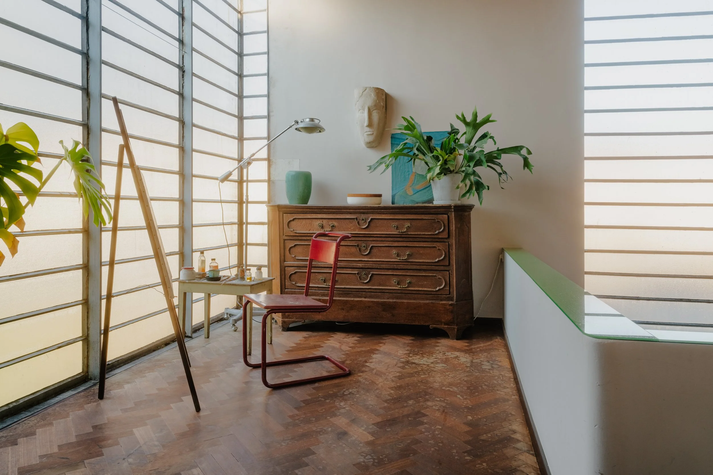 Interior of a bright room with large window blinds, a vintage wooden dresser with plants and art on top, a red chair, and art supplies on a small table.
