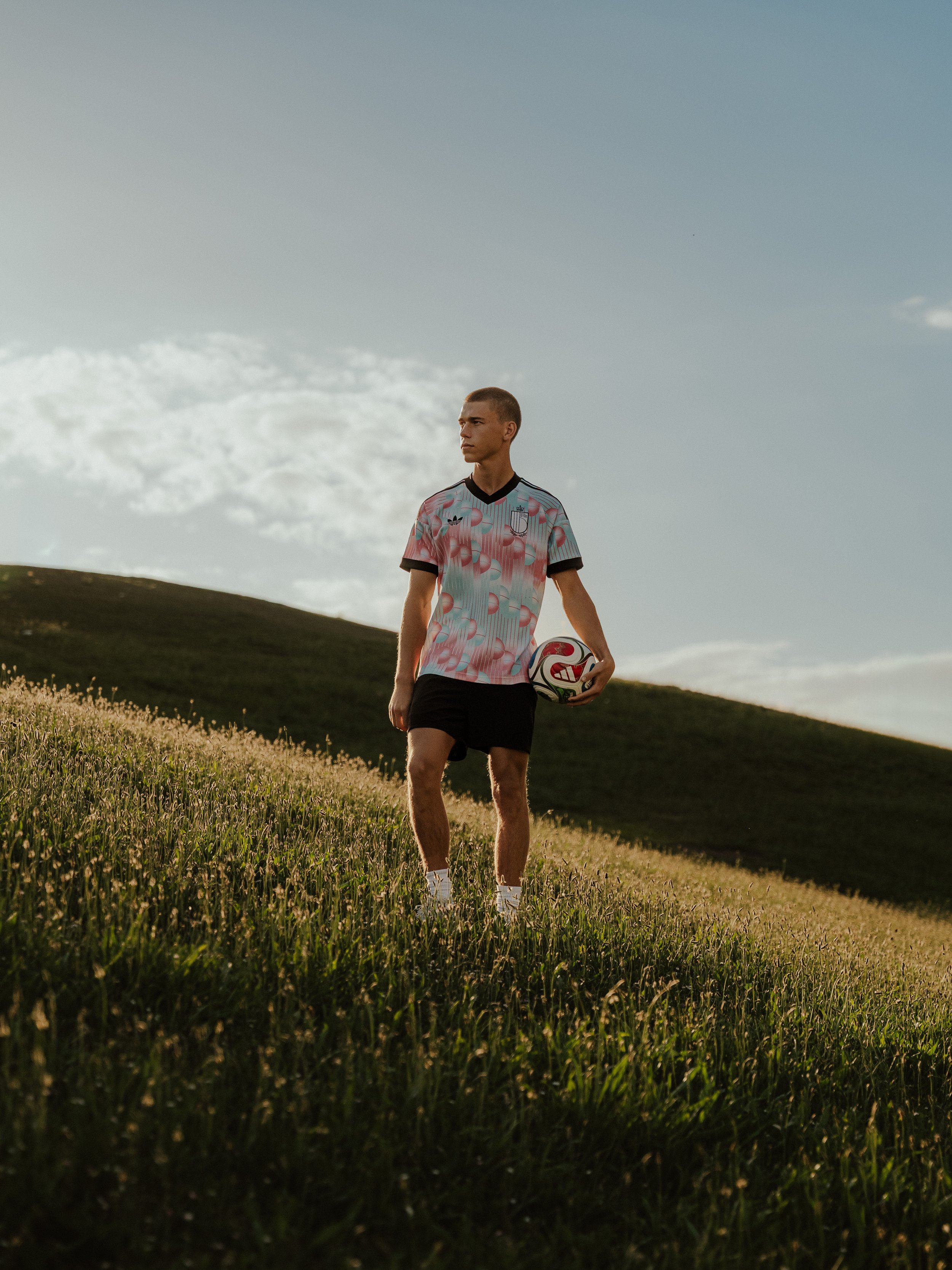 A young man stands outdoors in a grassy field, holding a soccer ball, with hills and a partly cloudy sky in the background.