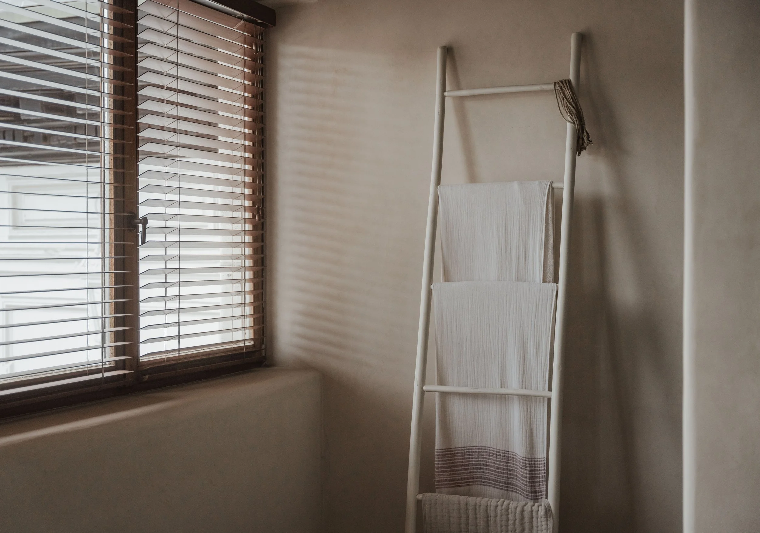 Interior scene with a window having wooden blinds, a white towel draped over a freestanding towel rack, and sunlight casting shadows through the blinds on the wall.