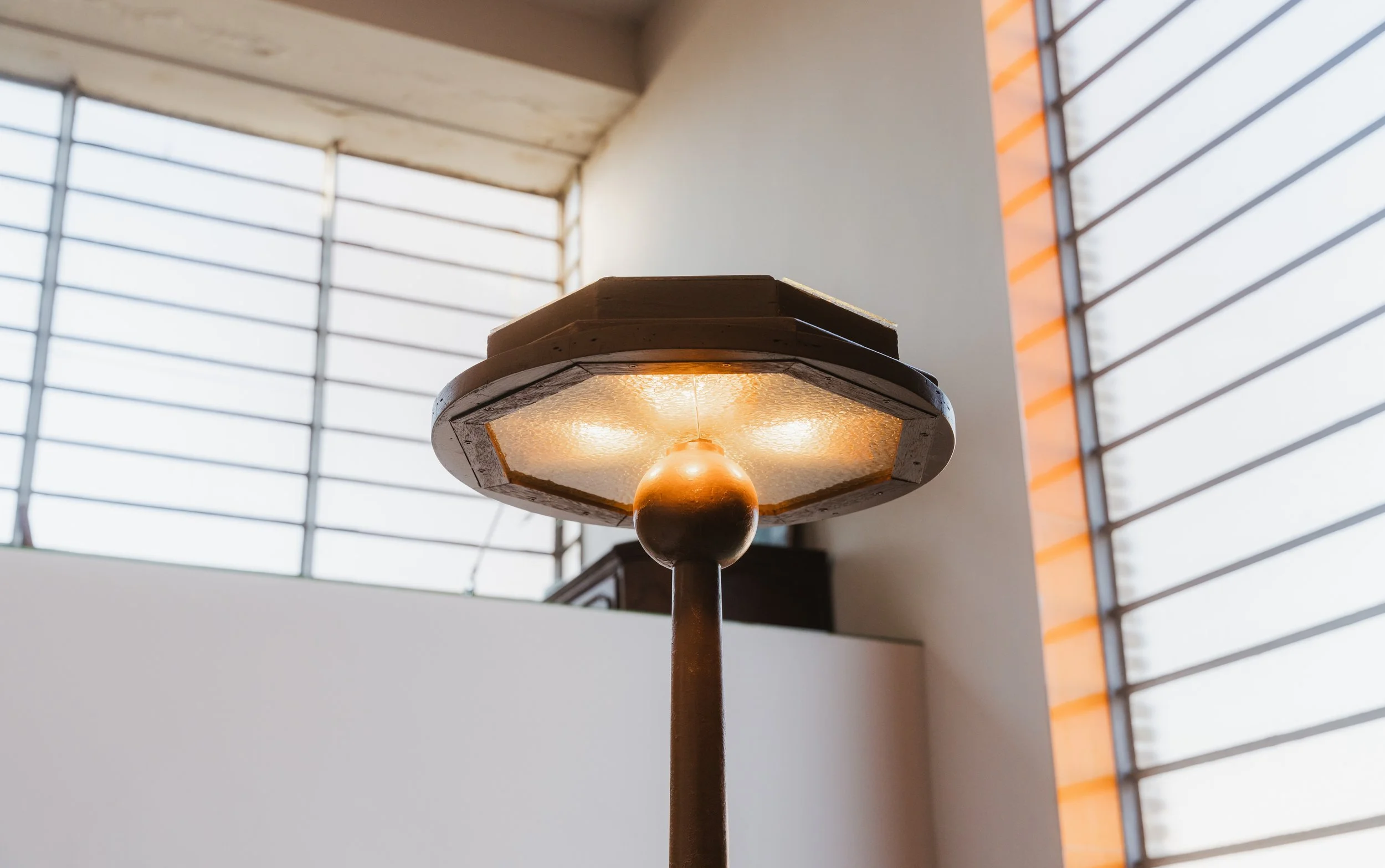A vintage-style standing lamp with a round glass or textured glass shade and a brown finial in an indoor setting with large window blinds.