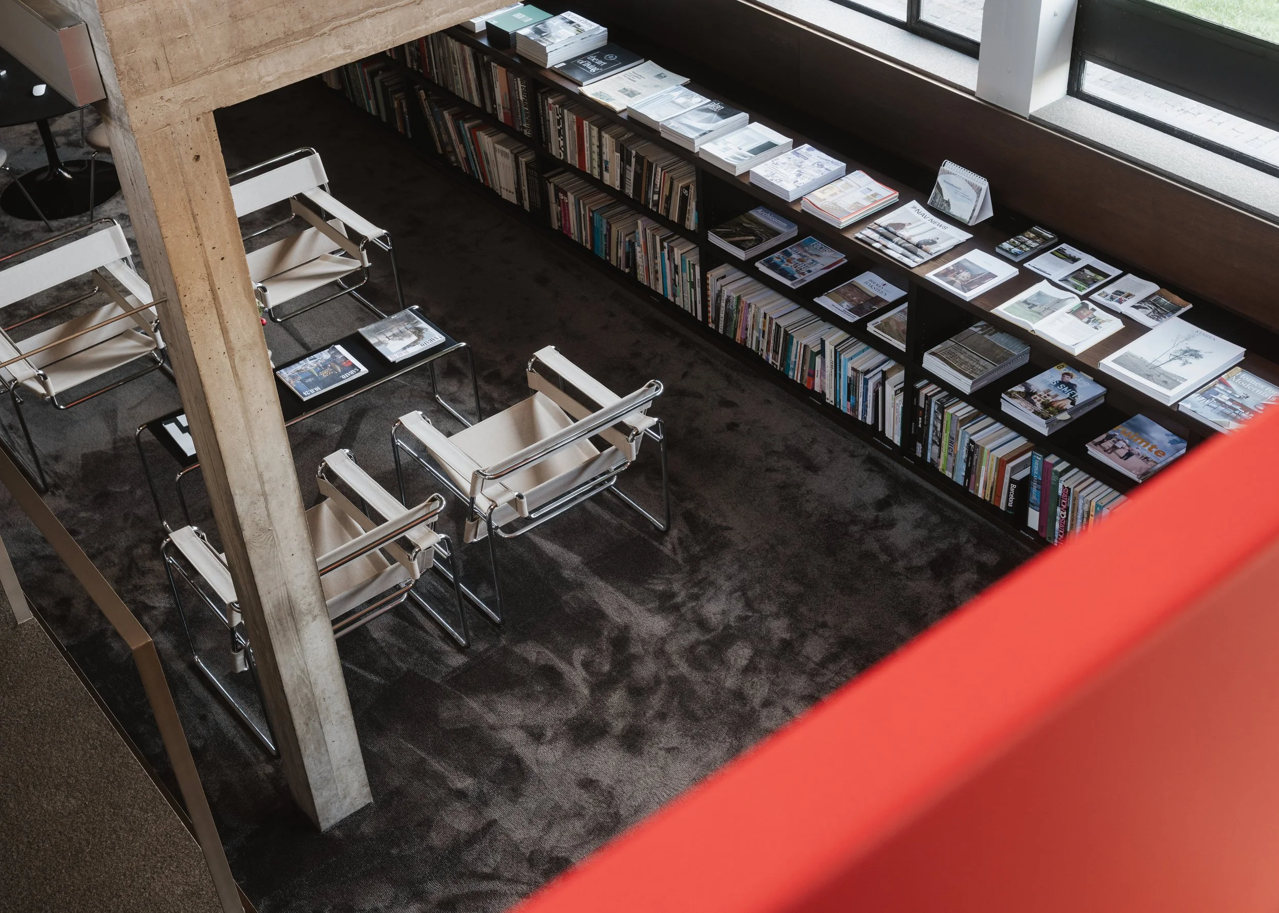 View of a modern library or bookstore interior with a black carpet, white chairs with metal frames, and shelves filled with books and magazines, seen from above near a large window.