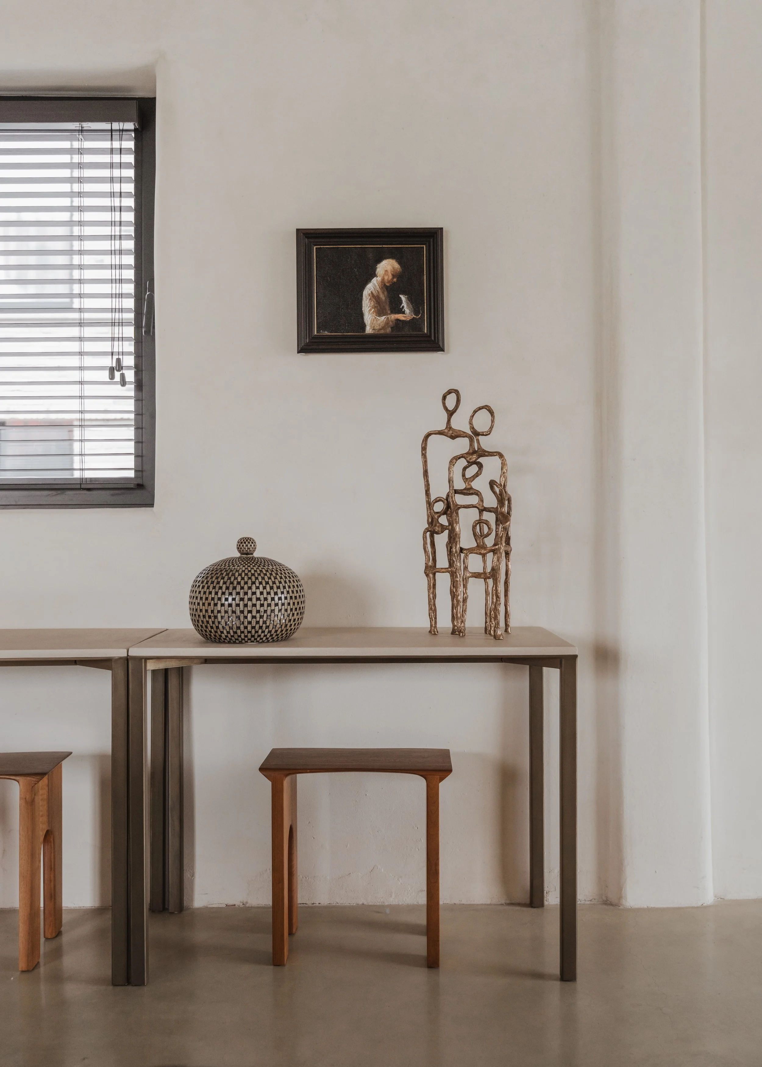 Interior room with a table, two small wooden stools, a decorative urn, a metal sculpture of abstract human figures, a framed painting of a person reading, and a window with metal blinds.