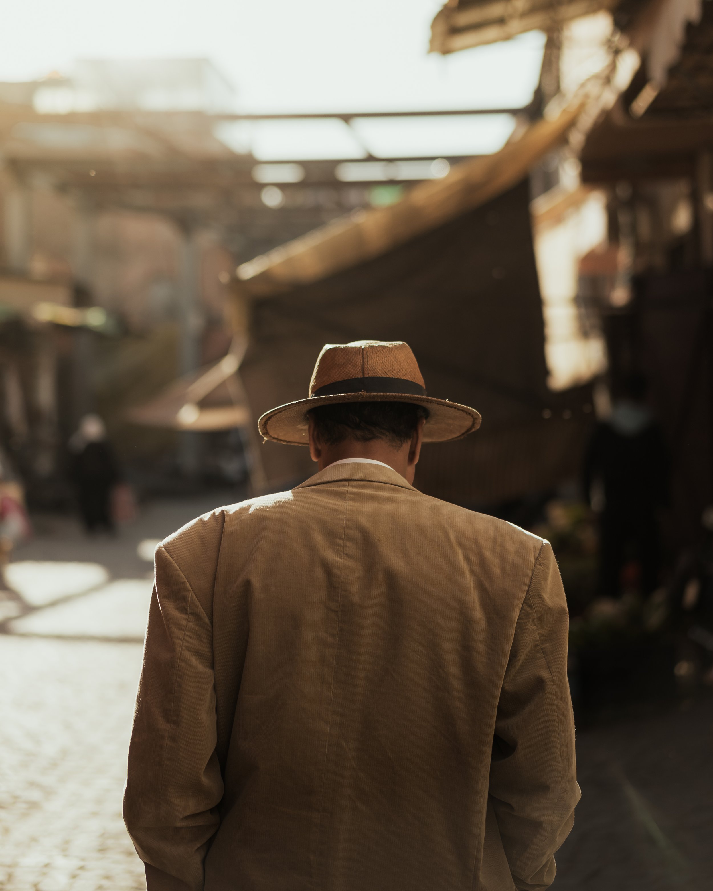 A man wearing a brown hat and beige jacket walking in an outdoor market during the daytime.
