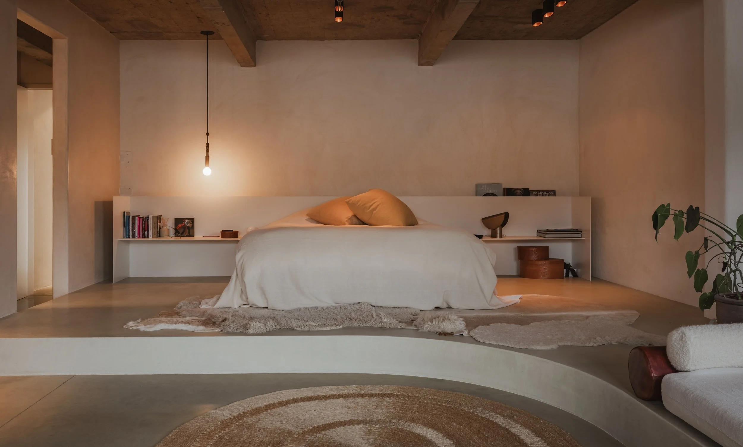 Minimalist bedroom with a low platform bed, beige bedding and pillows, bookshelf with books, decorative items, hanging light, and a potted plant.