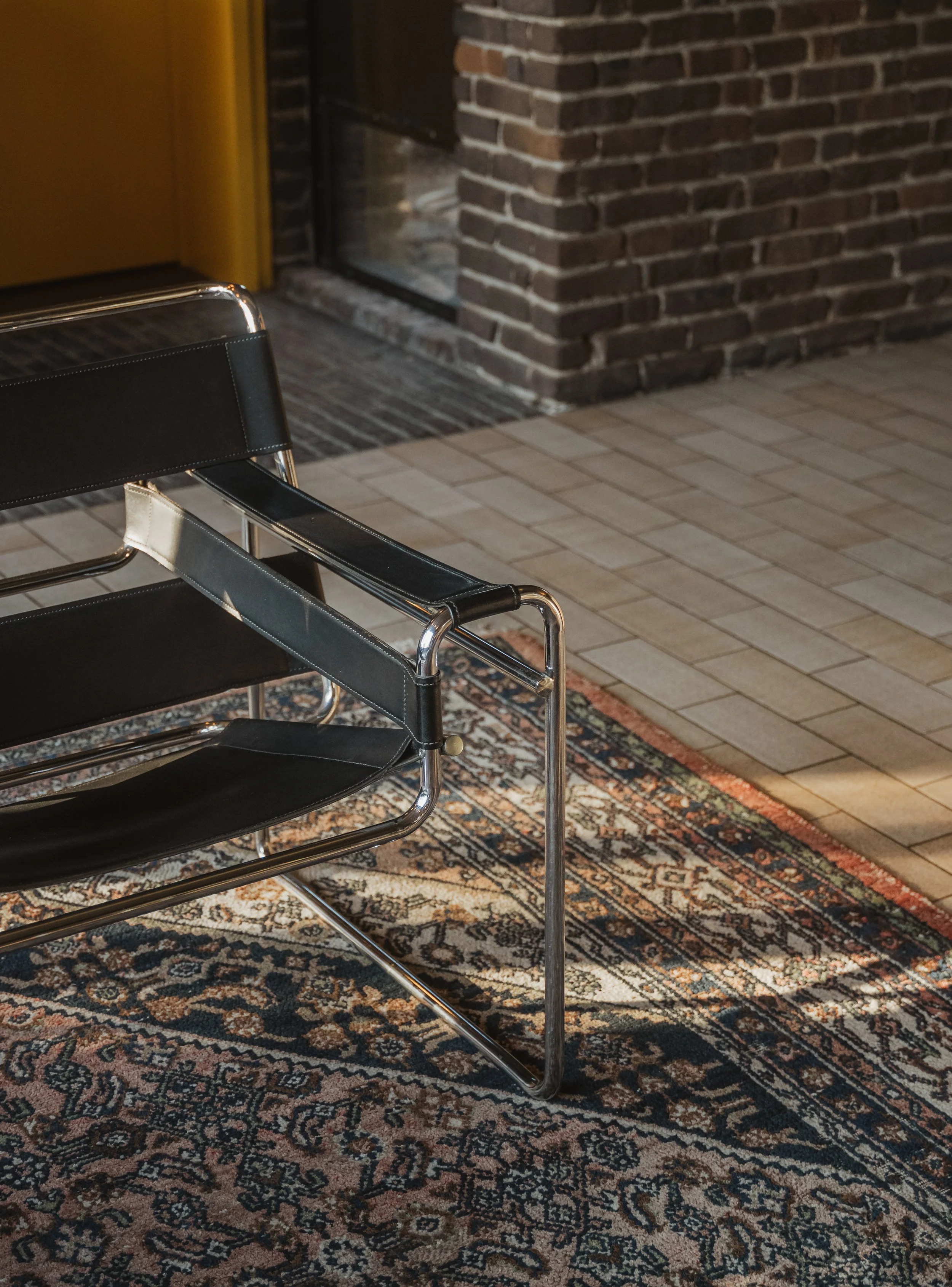A black and chrome lounge chair with a Persian rug underneath, situated in front of a brick wall and a glass door.
