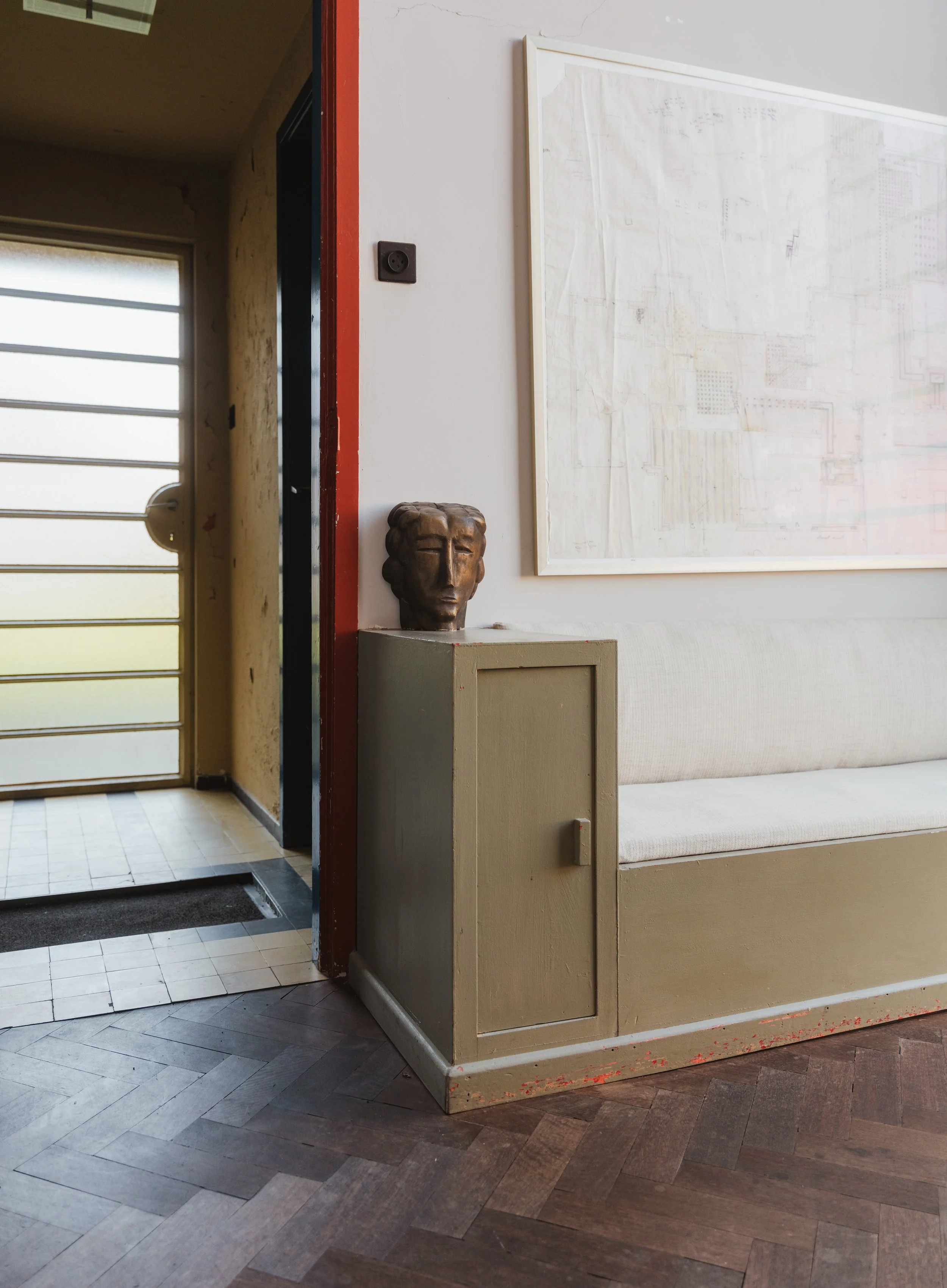 Interior corner with a beige bench, a small cabinet, a decorative face sculpture on the cabinet, a framed drawing on the wall, a partially visible glass door with horizontal stripes, and an outside door with a metal grid.