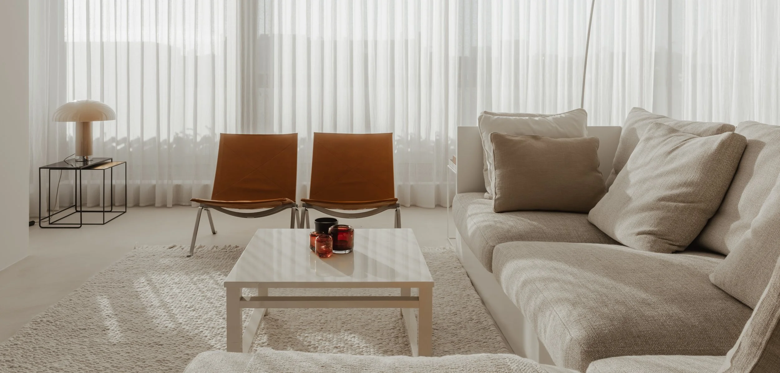 Bright living room with beige sofa, white coffee table, two brown chairs, a black side table with a white lamp, sheer white curtains, and neutral decor.