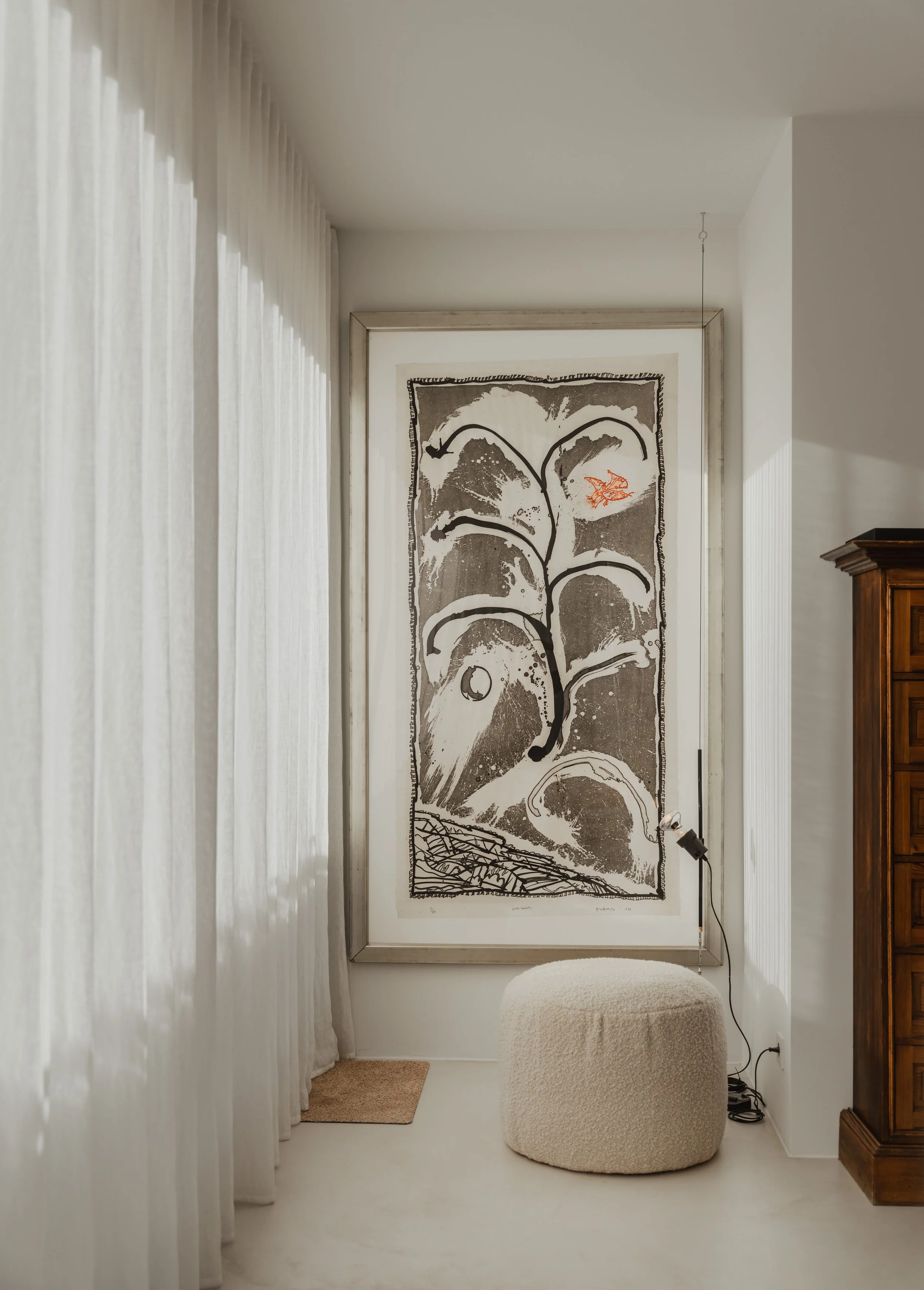 Minimalist interior with large abstract black and white artwork on the wall, a beige textured ottoman, sheer white curtains, a small beige rug, a wooden cabinet, and a floor lamp with a clip on light