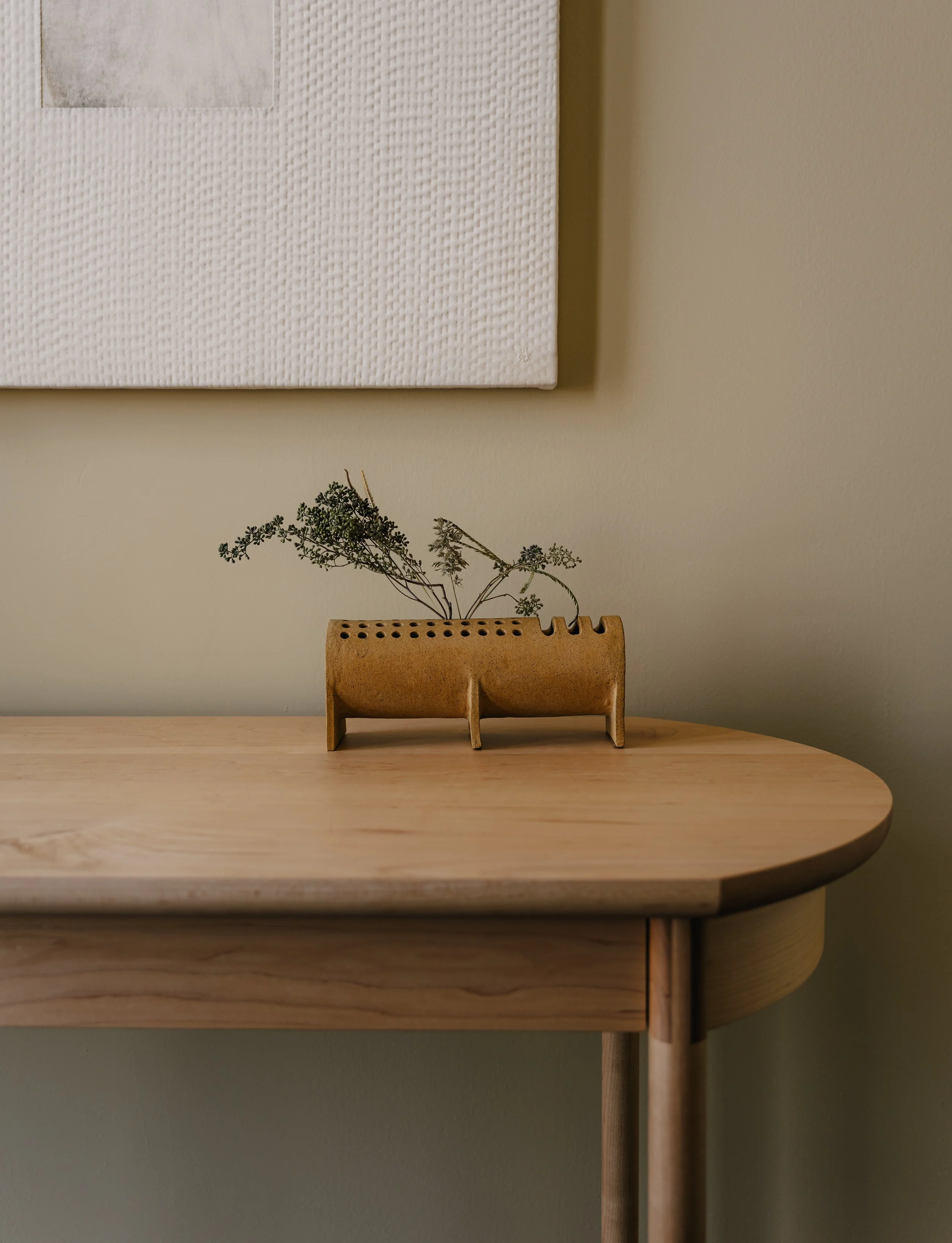 Close-up of a light-colored wooden table with a small, rectangular, brown wooden organizer holding dried plant stems, against a beige wall and a canvas with textured white and gray artwork.